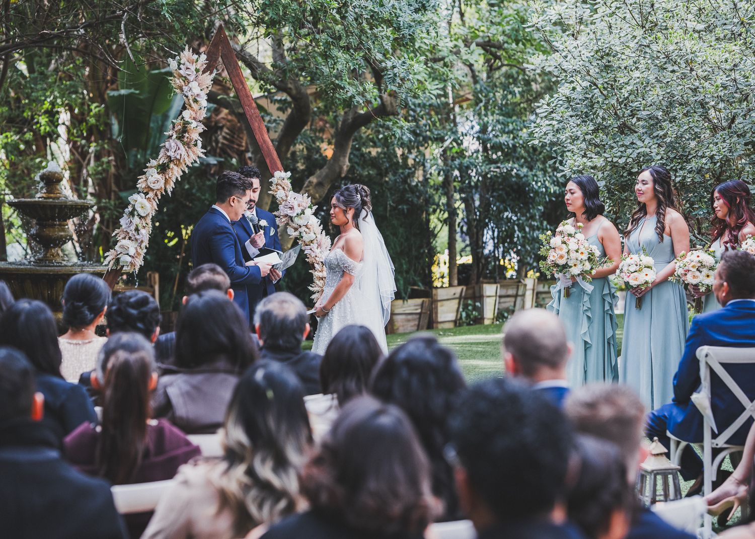 Outdoor garden wedding ceremony with guests seated and floral arch decorations.
