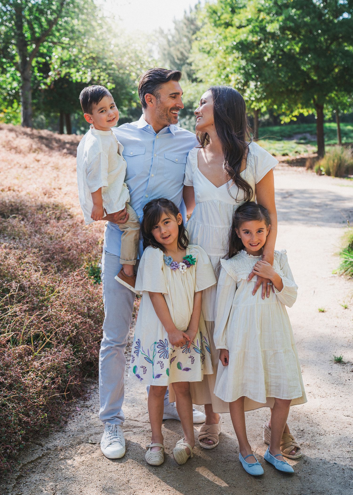 Family spending time together on a sunlit garden path wearing coordinated white and light blue outfits.
