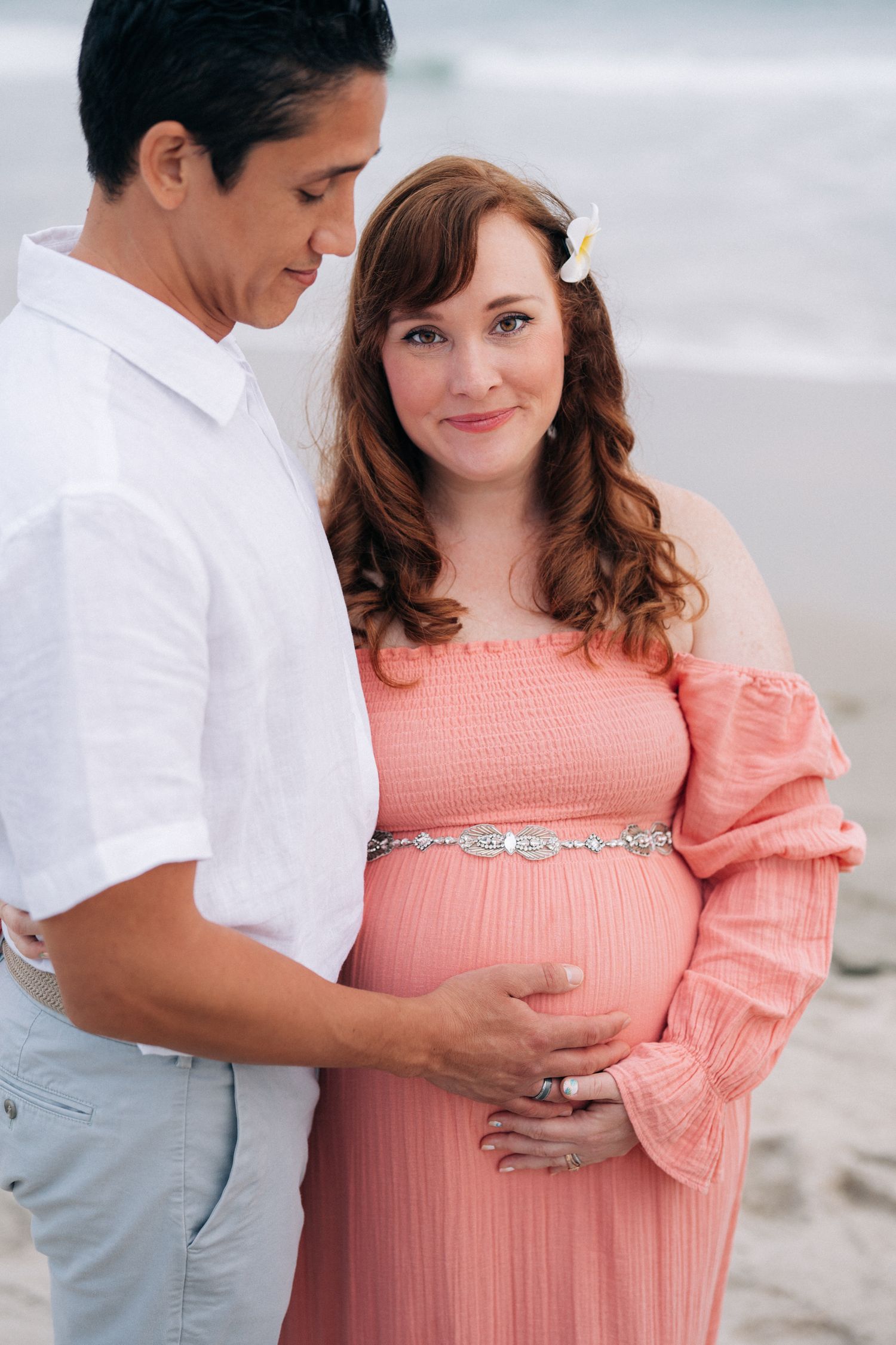 A maternity photoshoot featuring a couple in white and coral attire on a cloudy beach.