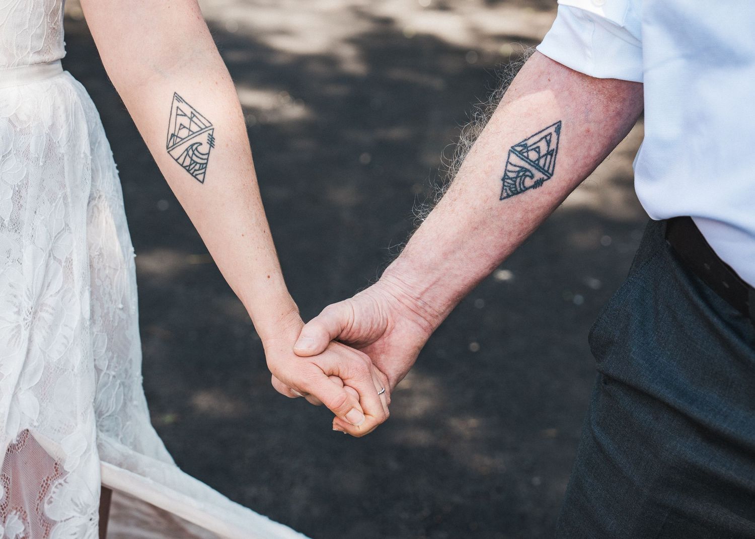 Two people holding hands showcase matching geometric diamond shape tattoos on their forearms in a romantic gesture.