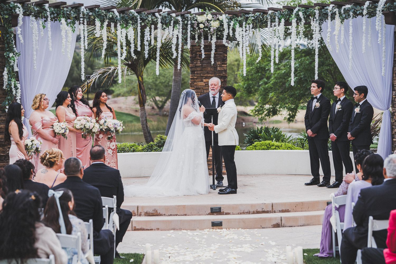 An elegant outdoor wedding ceremony takes place under white streamers hanging from trees with bridal party and guests present.