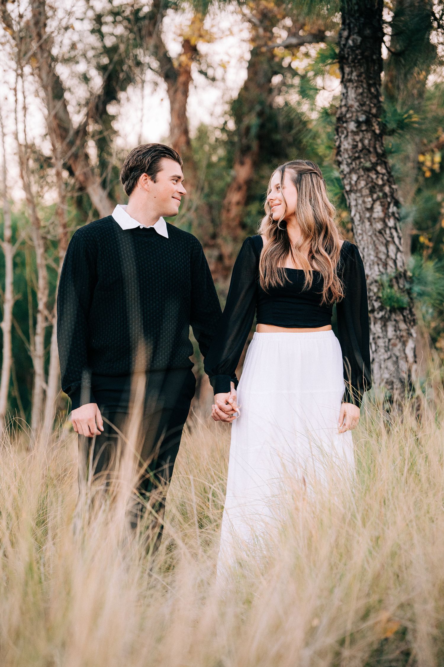 Couple in black and white attire poses intimately in tall grass field with trees in background during sunset.