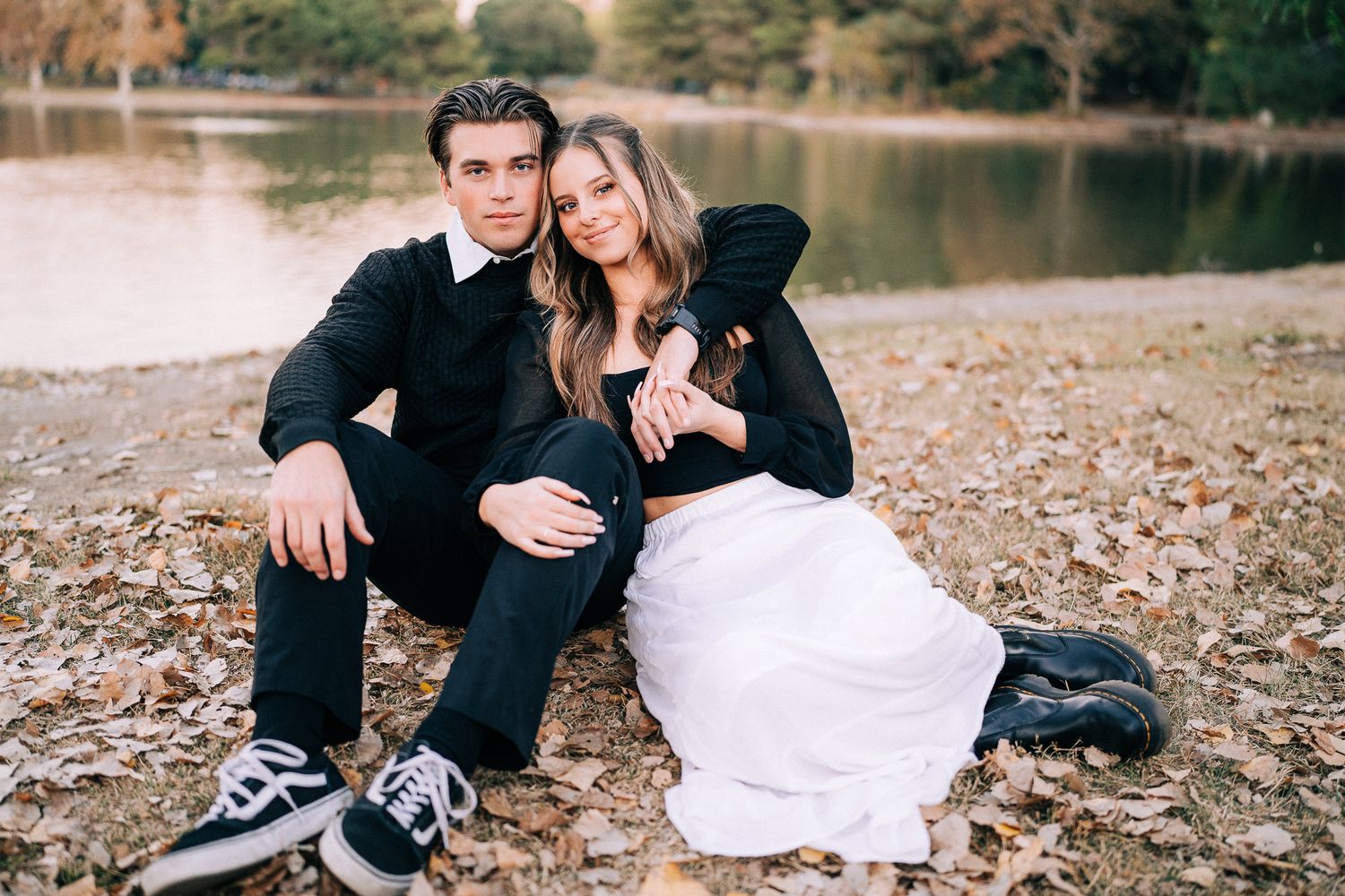 Happy couple laying in autumn leaves enjoying a romantic engagement photo session near a lake.