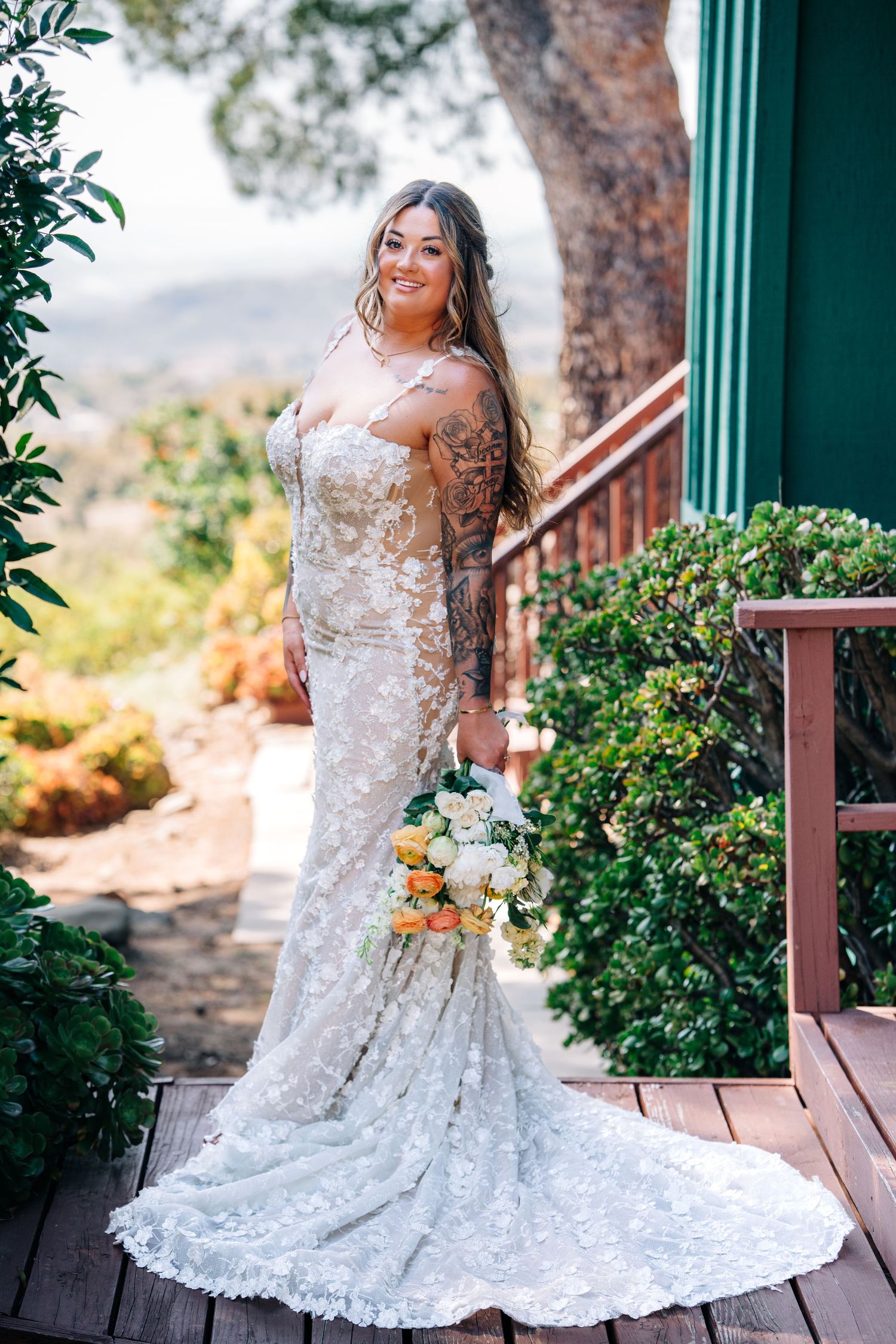 A stunning lace wedding dress with a long train is showcased on a wooden deck surrounded by greenery and mountain views.