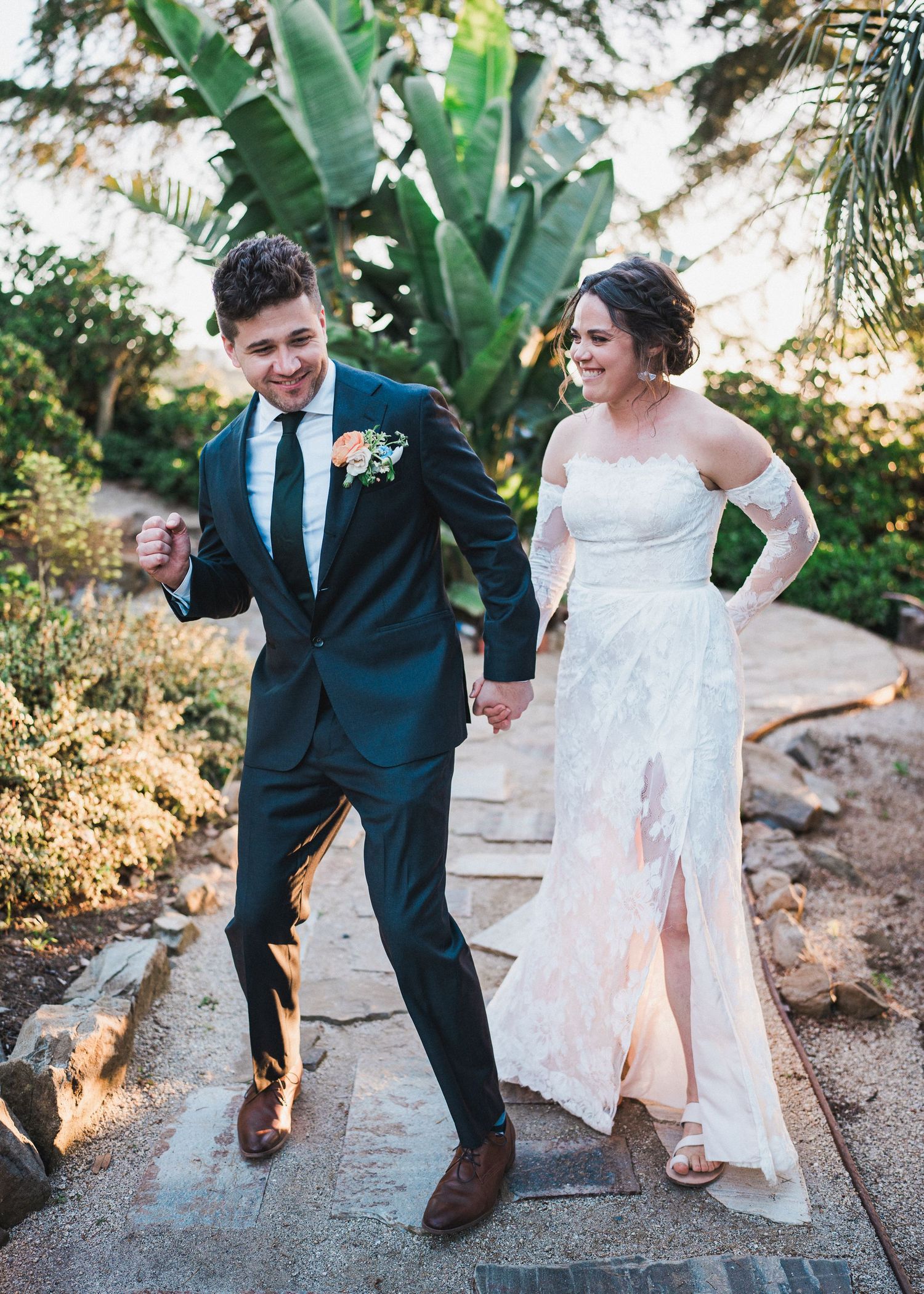 A newlywed couple laughs while walking down stone steps surrounded by tropical plants.