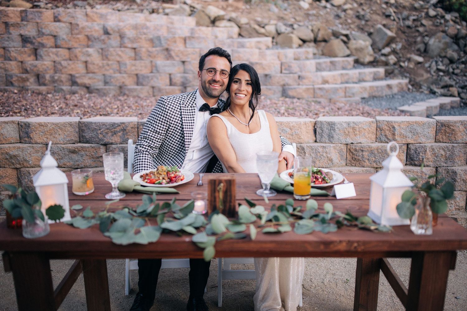 A newlywed couple poses behind their rustic wedding reception table decorated with candles and refreshments.