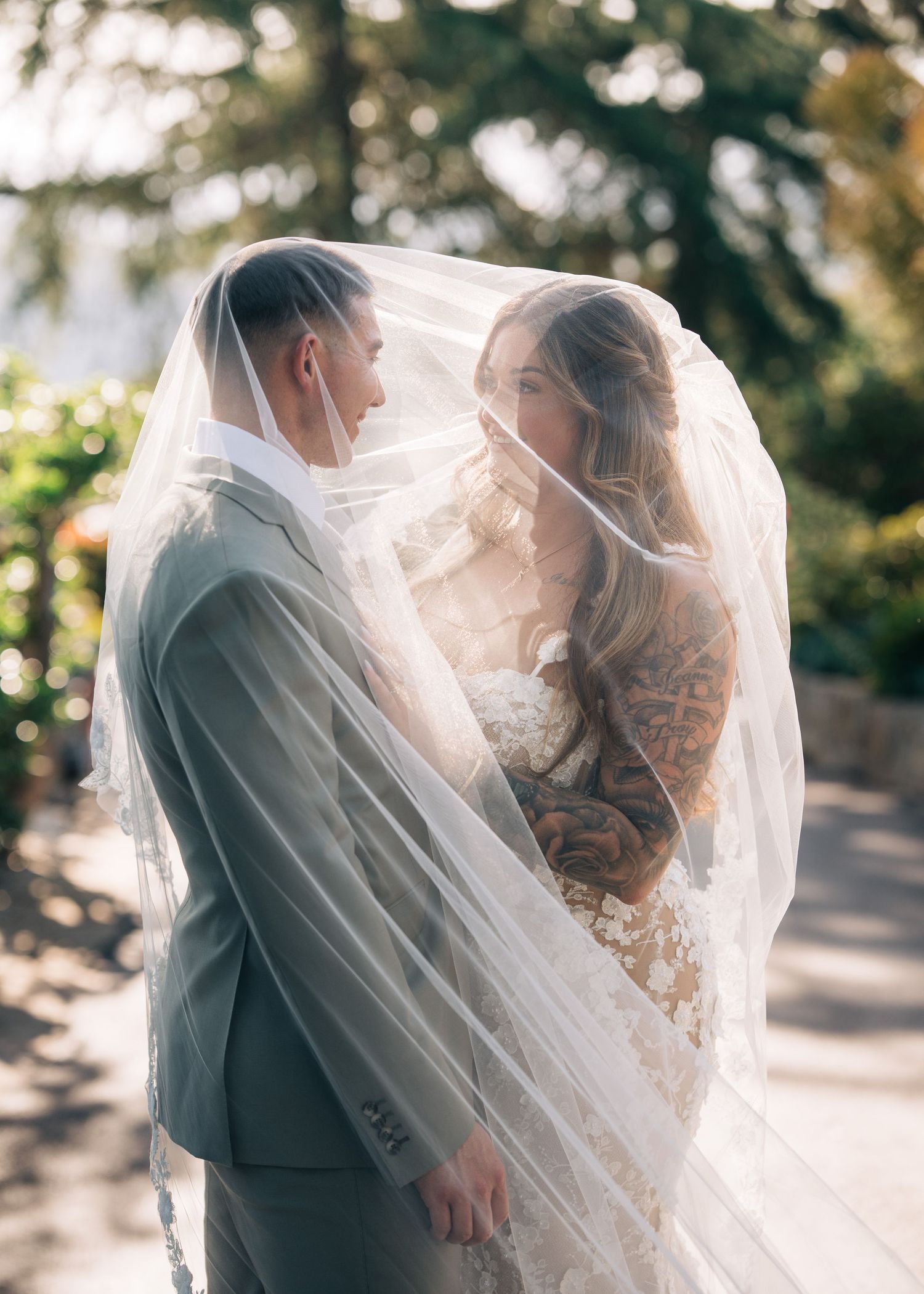 A romantic wedding moment captured as a bridal veil flows around the couple in soft sunlight.