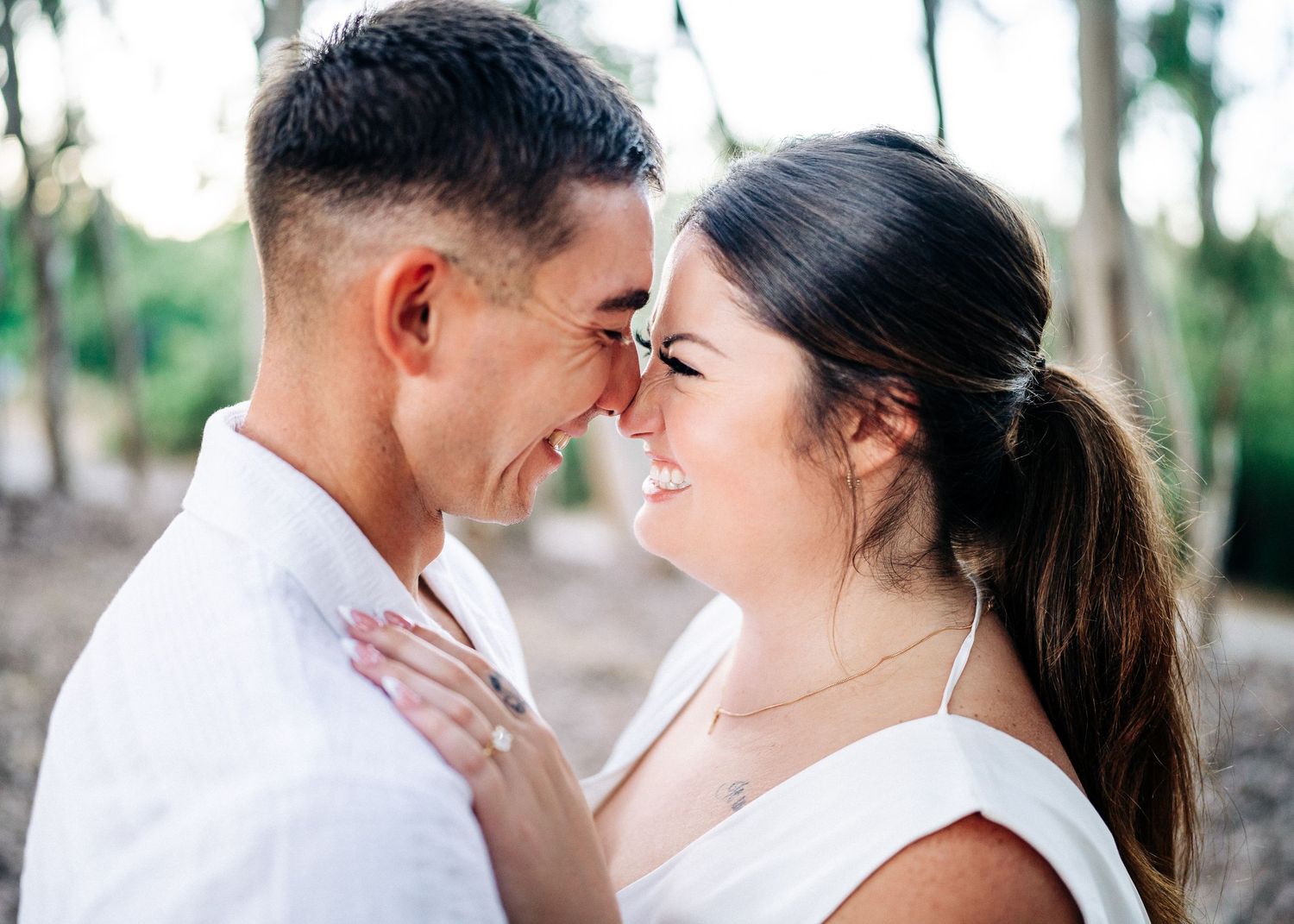 A romantic moment between a couple in white attire sharing a tender close embrace outdoors in a wooded setting.