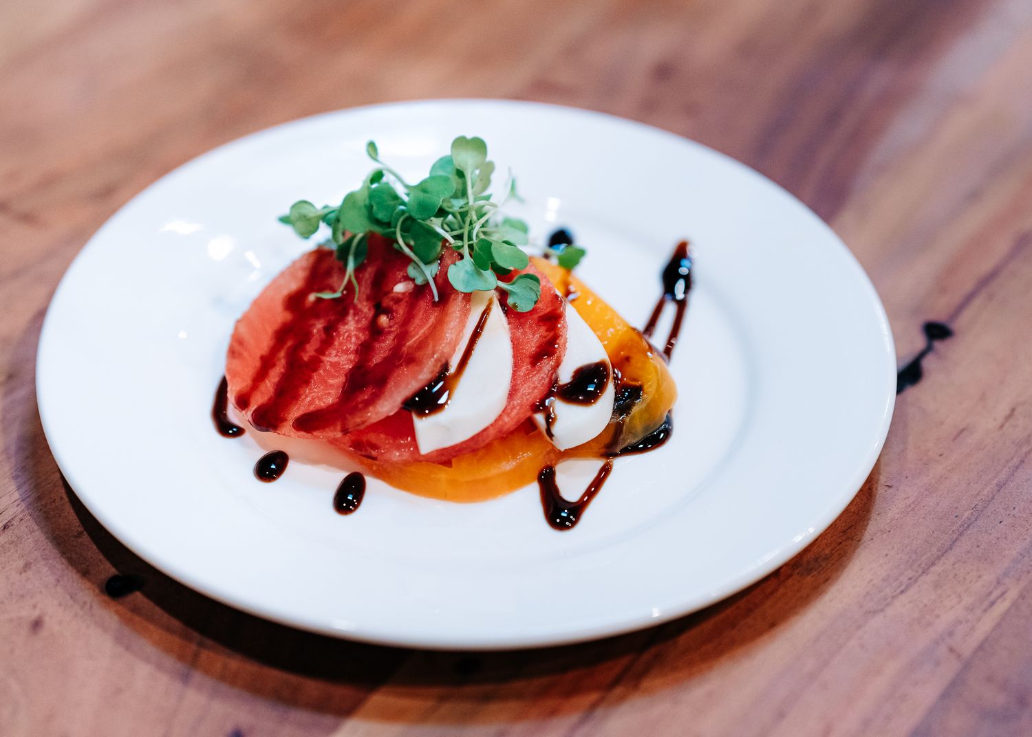 Elegant plated tomato salad with microgreens drizzled with balsamic glaze served on a white plate on wooden surface.