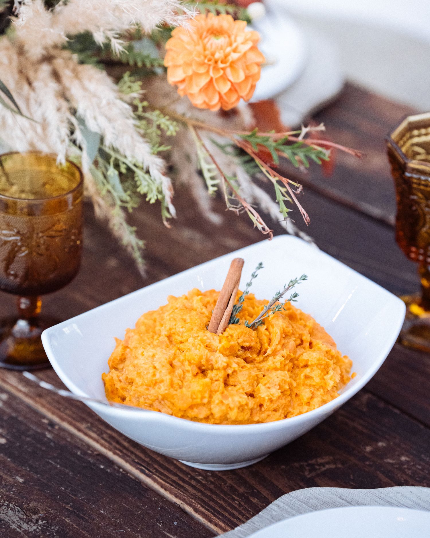 Bowl of vibrant orange carrot puree garnished with herbs and served with rustic glassware and floral decorations.