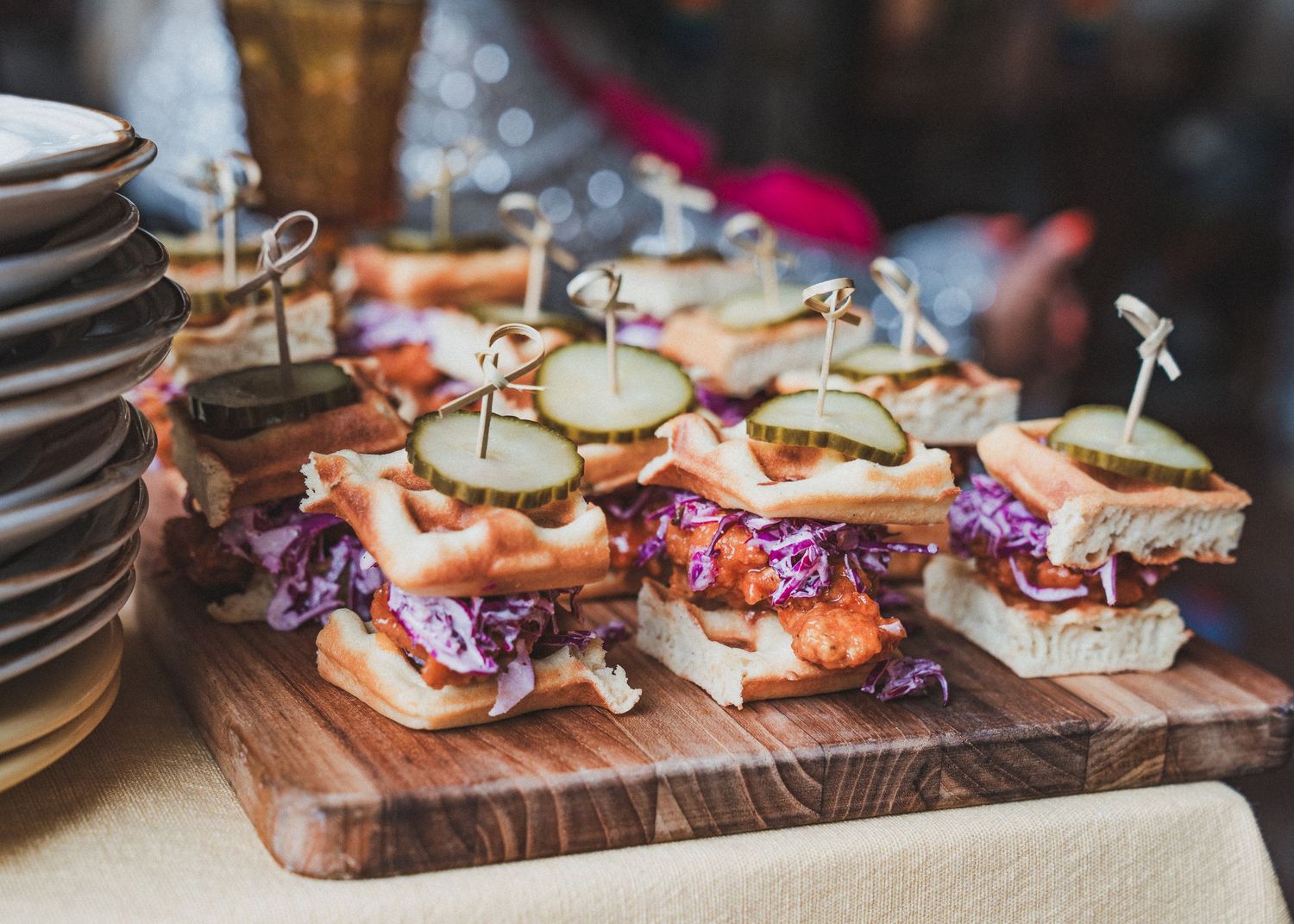 Mini sandwiches with colorful toppings and garnishes arranged on a rustic wooden serving board at a catering event.