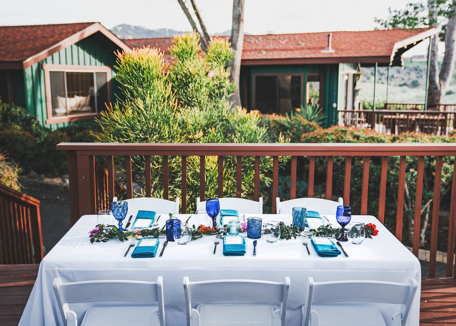 A table set with blue and white decor on a wooden deck patio with rustic cabins in the background.
