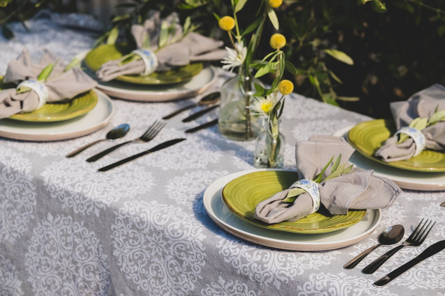 Elegant outdoor table setting with green plates, linen napkins, black cutlery, and yellow floral accents on a white lace tablecloth.