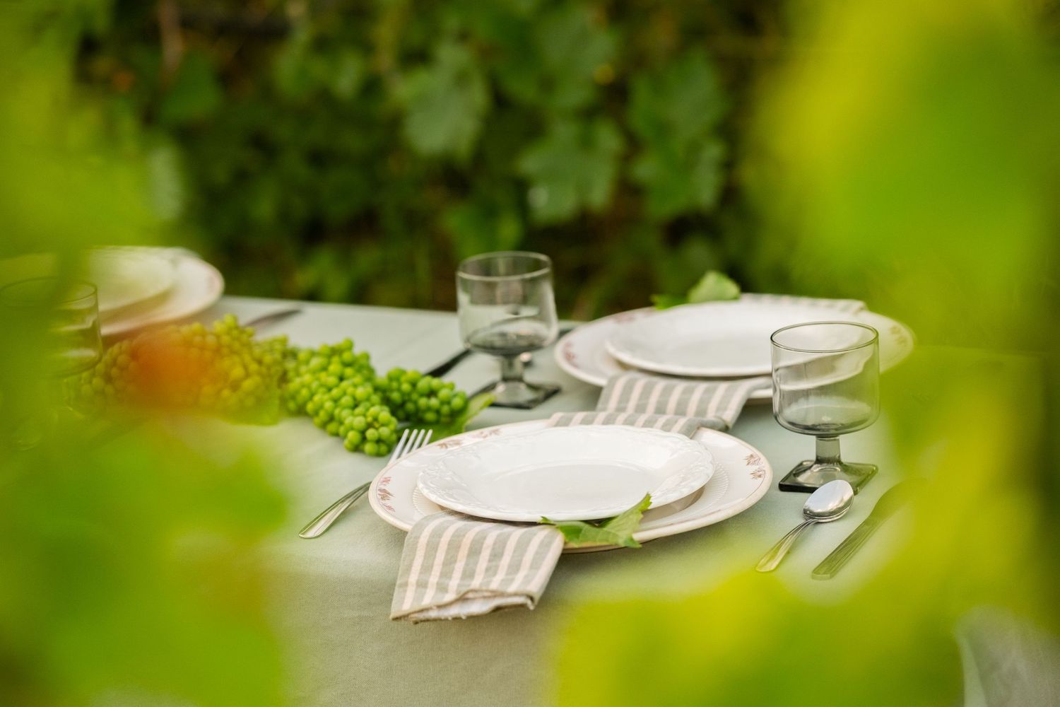 Elegant outdoor table setting with white plates, glassware, and green floral accents on a linen tablecloth.