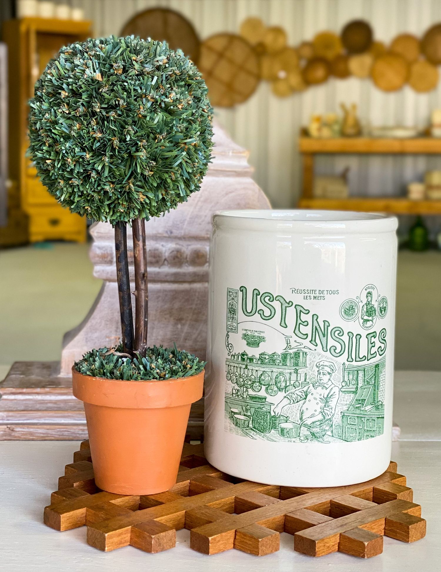 Decorative green topiary tree and white utensil holder displayed on wooden trivet with farmhouse decor.