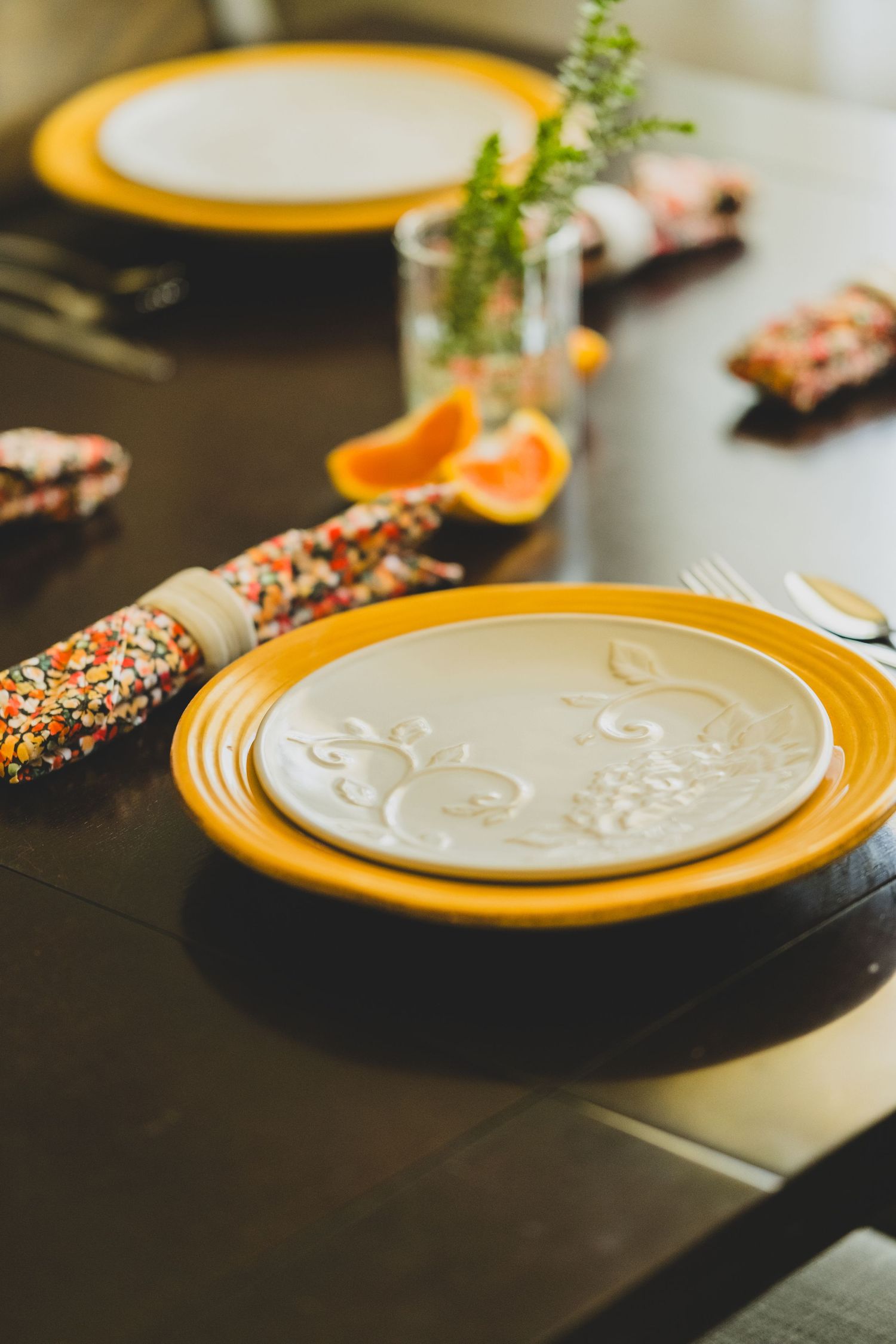 Elegant table setting with yellow ceramic plates, embossed white dinner plate, colorful napkin rings, and fresh orange slices.