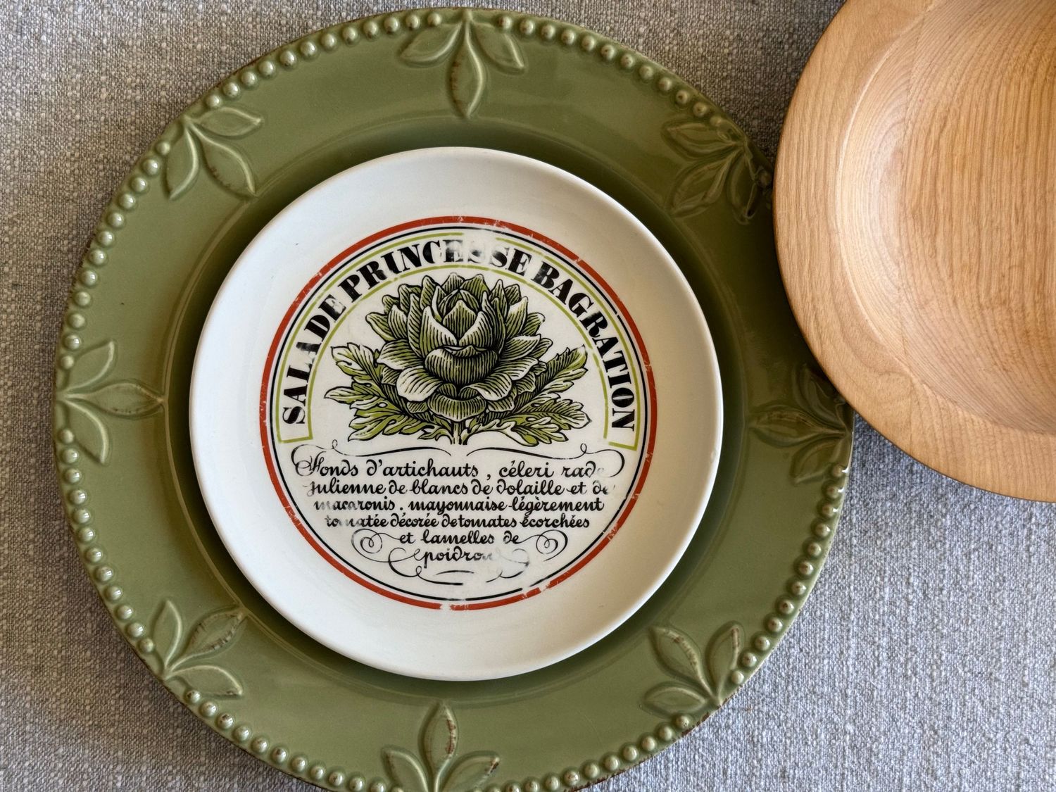 Vintage French ceramic salad plate with artichoke illustration on green embossed charger beside a wooden bowl.