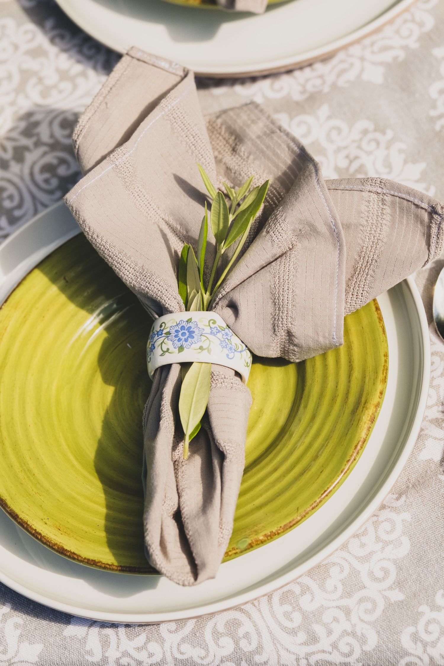 Elegant table setting with green plate, beige linen napkin, floral napkin ring, and olive branch sprig on white tablecloth.