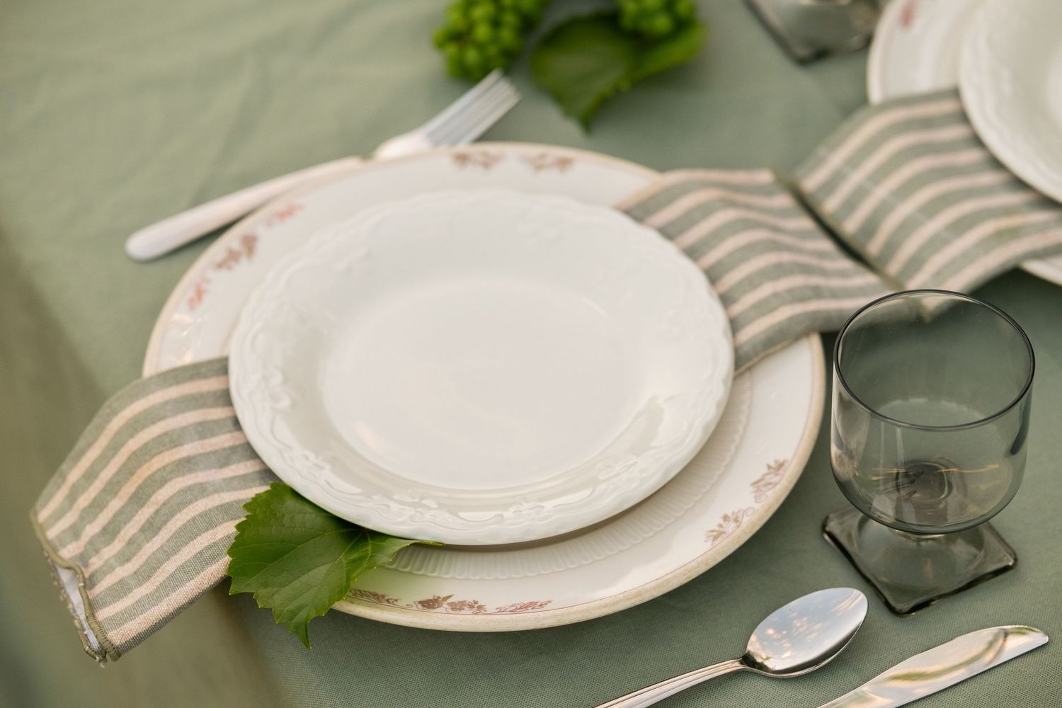 Elegant place setting with white plates, striped linen napkin, silverware, and a glass on a sage green tablecloth.