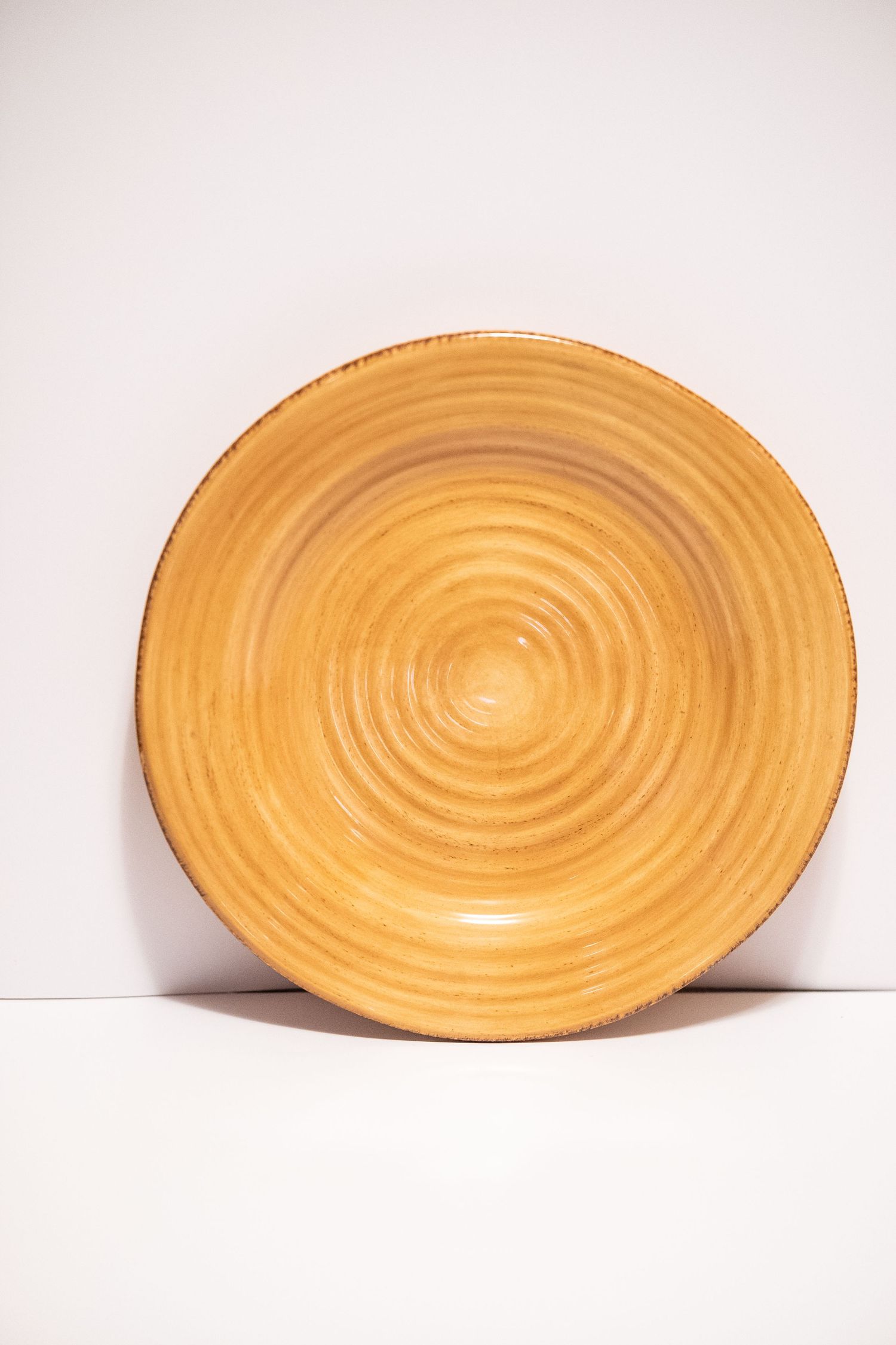 A round ceramic plate with concentric circle pattern in warm yellow-brown glaze, leaning against a white background.