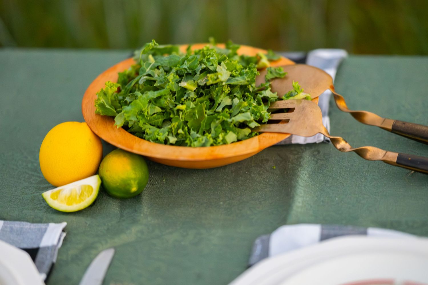 Fresh green herb salad in wooden bowl with lemon and lime on green table with serving utensils.