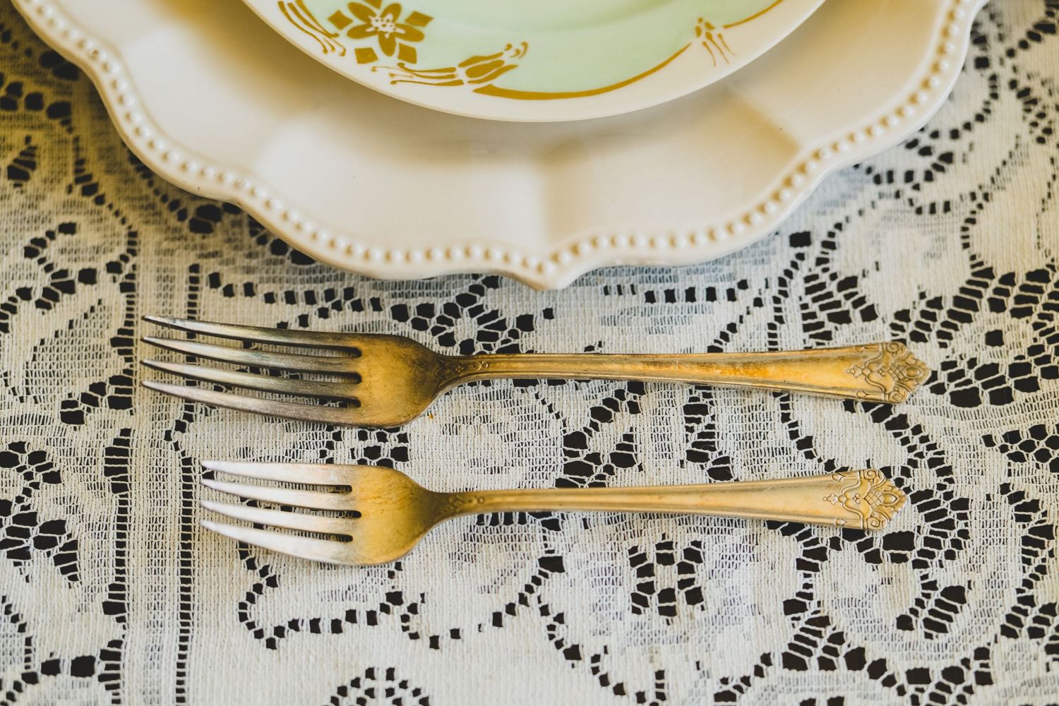 Two vintage forks, one gold and one silver, placed on a lace tablecloth beside an elegant floral plate.