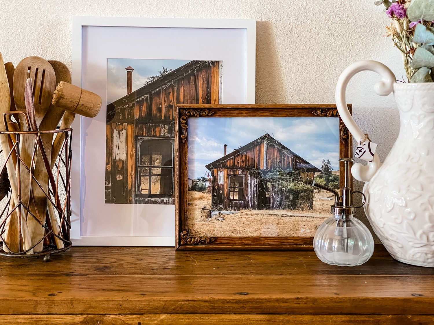 Framed rustic barn photos displayed on wooden surface with vintage white pitcher and wooden utensils.