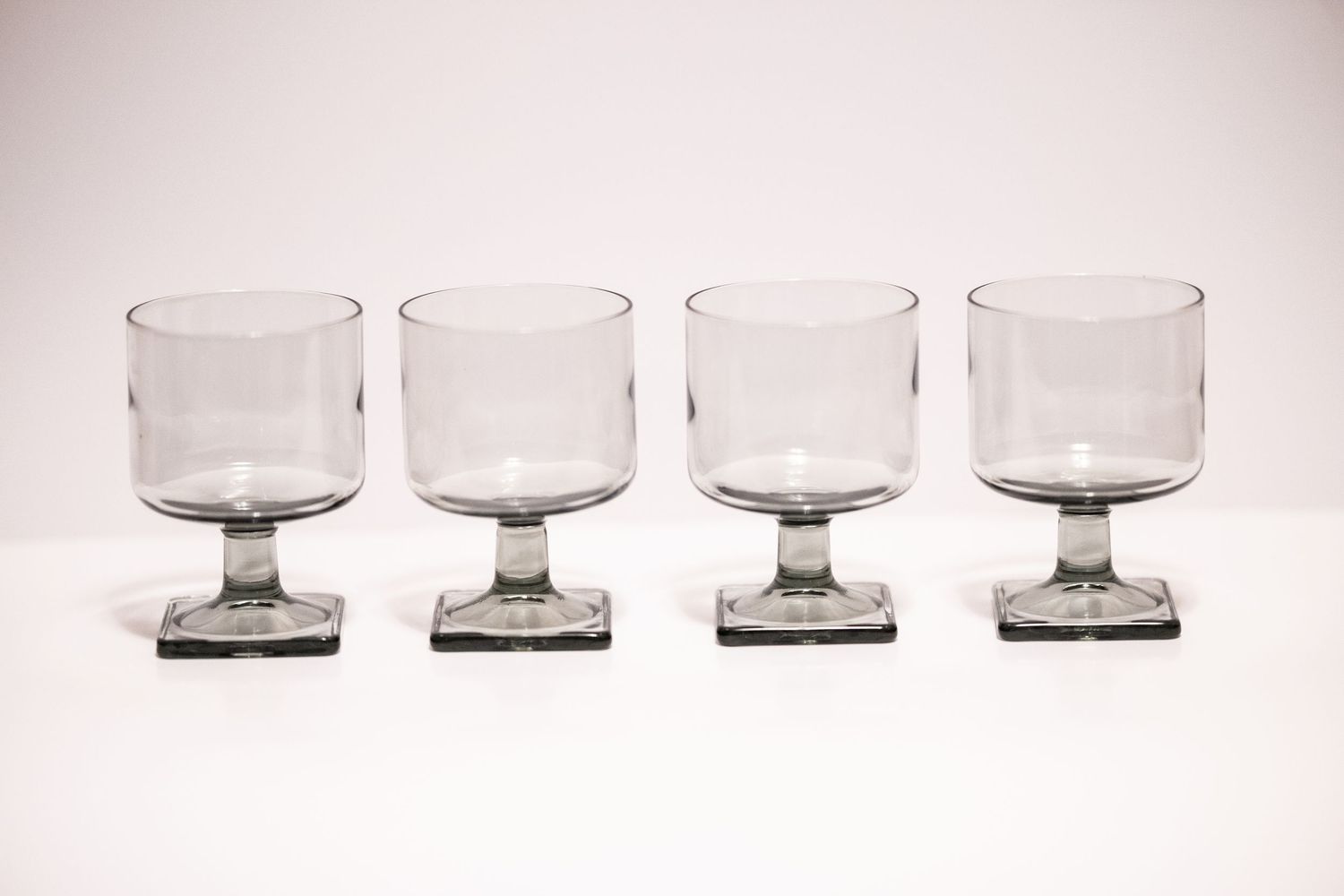 Set of four smoky gray glass goblets with square bases arranged in a row on a white background.
