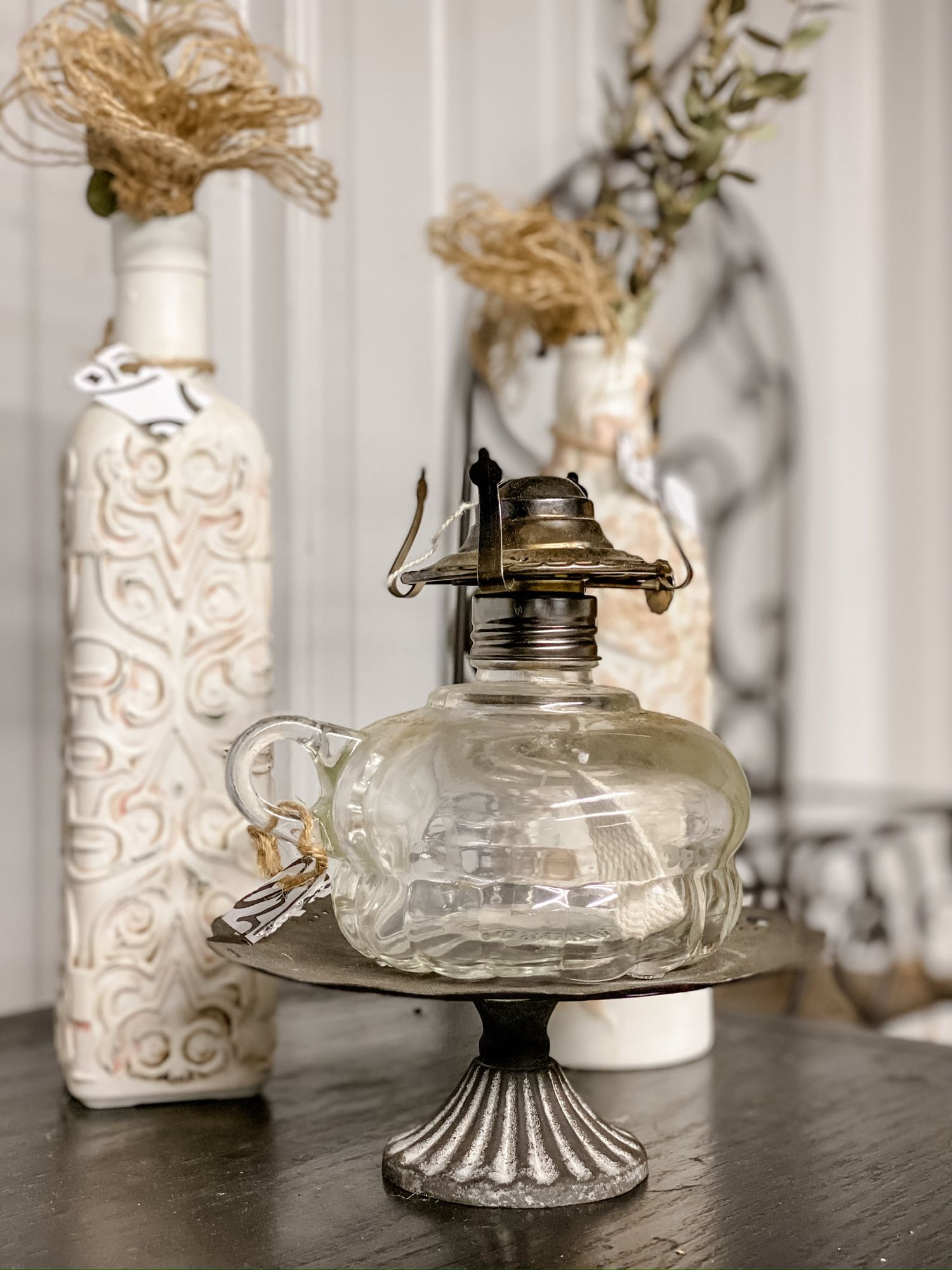 Antique glass oil lamp on silver pedestal with decorative white bottles in background.