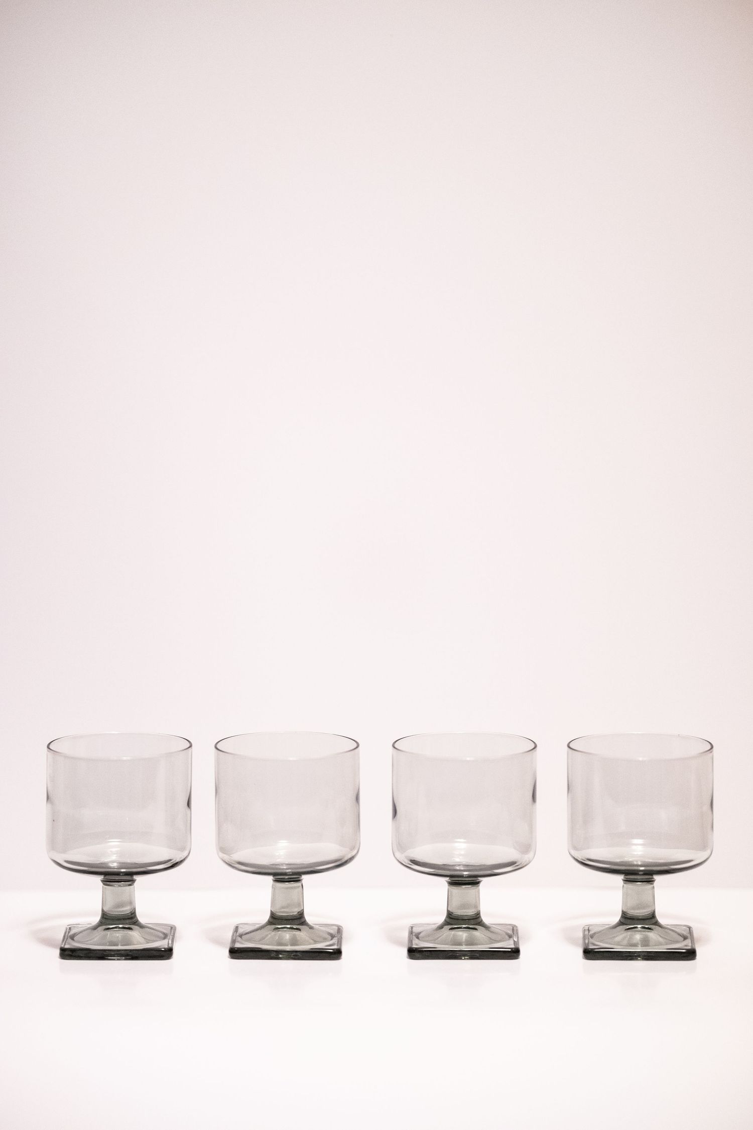 Four empty smoked glass footed tumblers with square bases arranged in a row on a white surface.