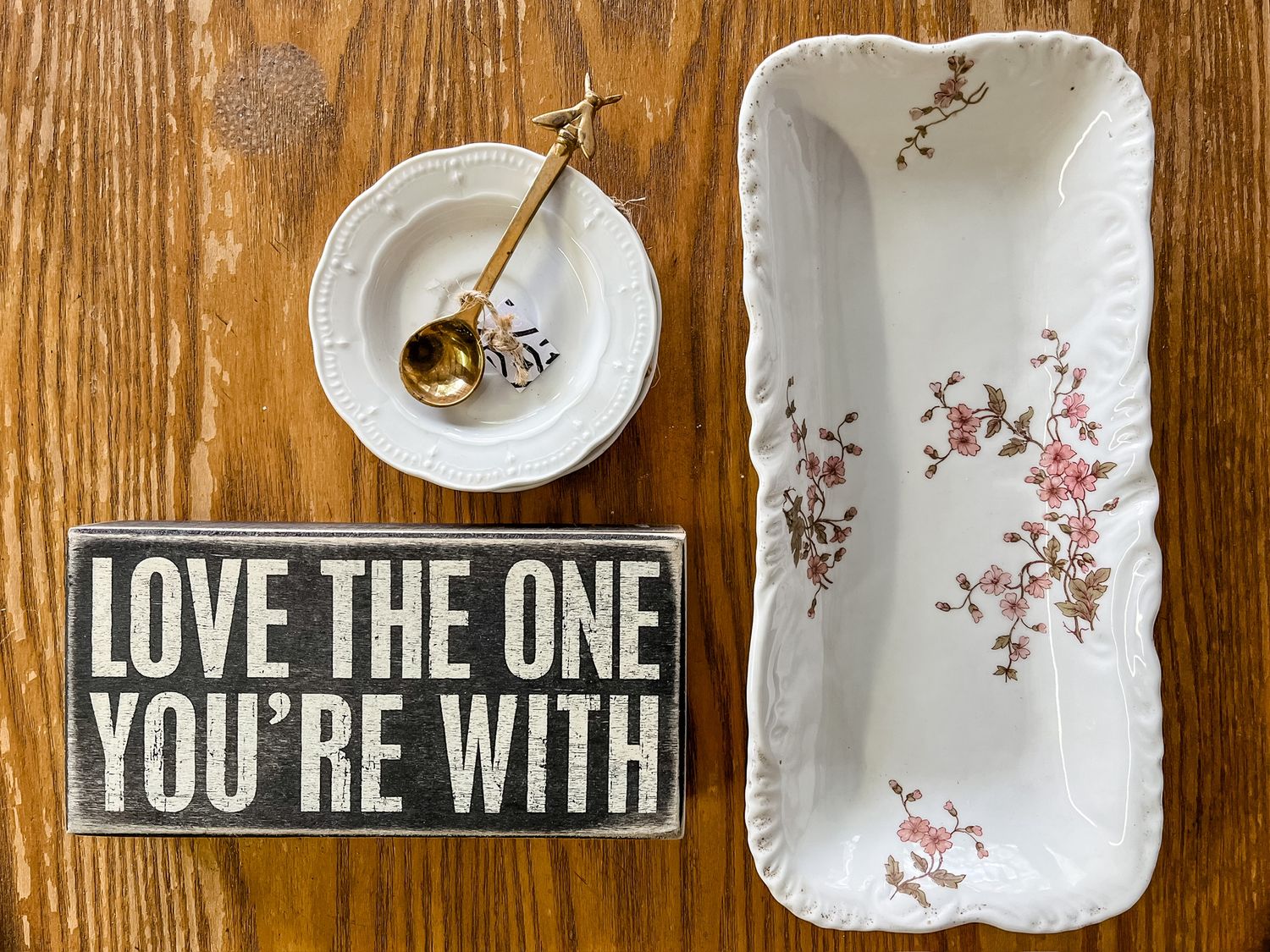 Flatlay of ceramic dishes with 'Love the one you're with' sign on wooden table surface.
