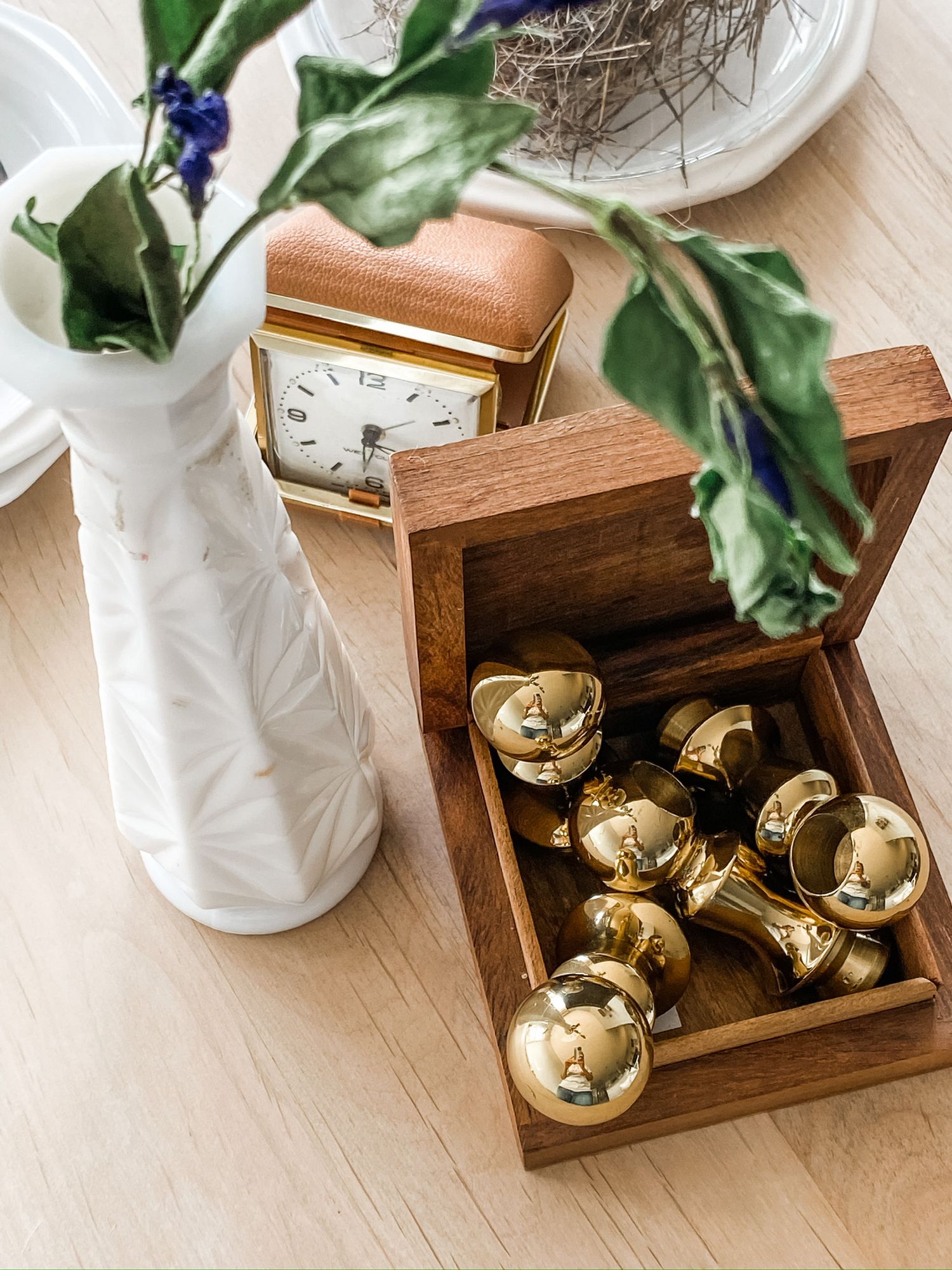 White ceramic vase with greenery next to wooden box filled with gold bells on light surface.