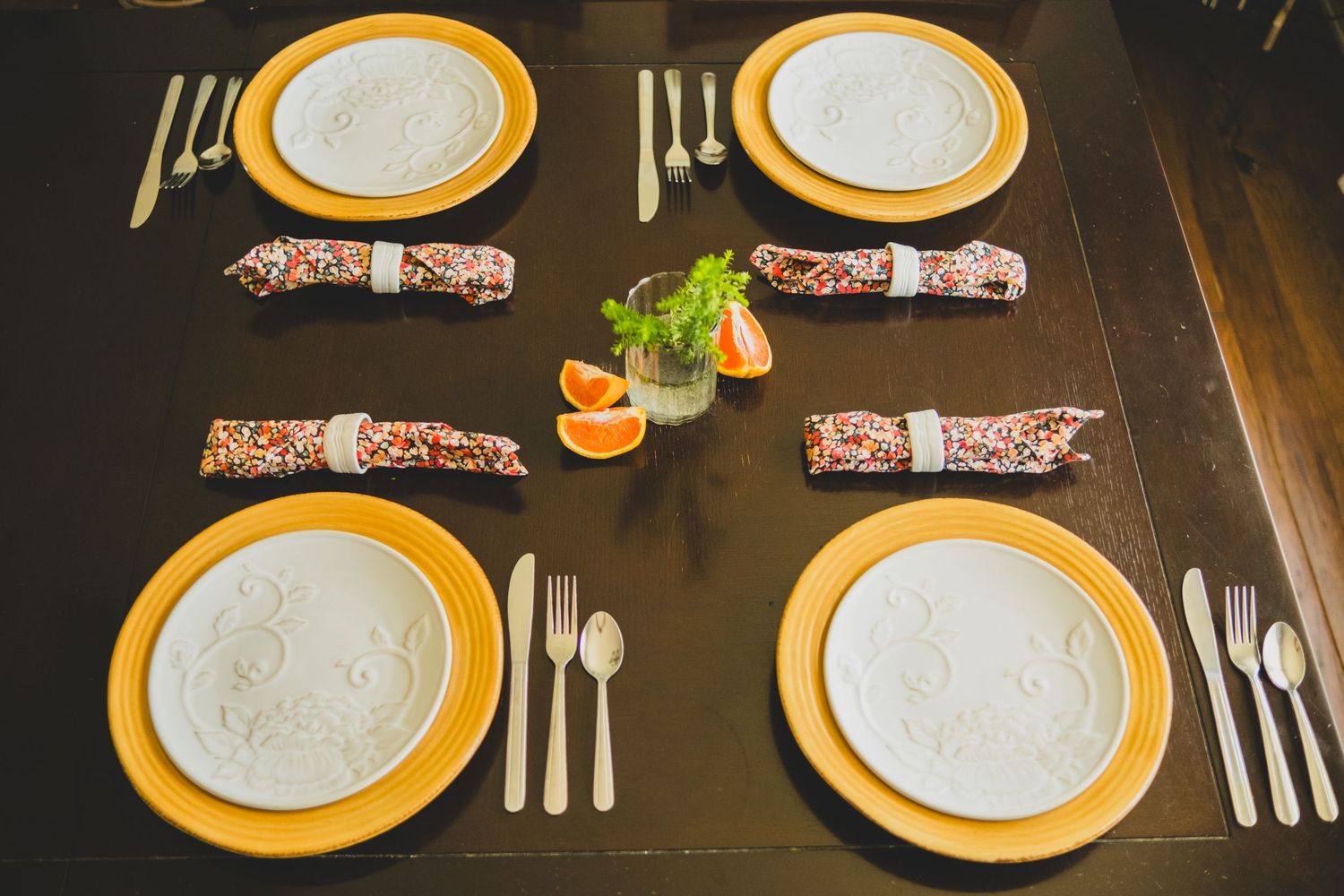 Elegant table setting with yellow-rimmed white plates, rolled floral napkins, silverware, and orange slices on a dark wood table.