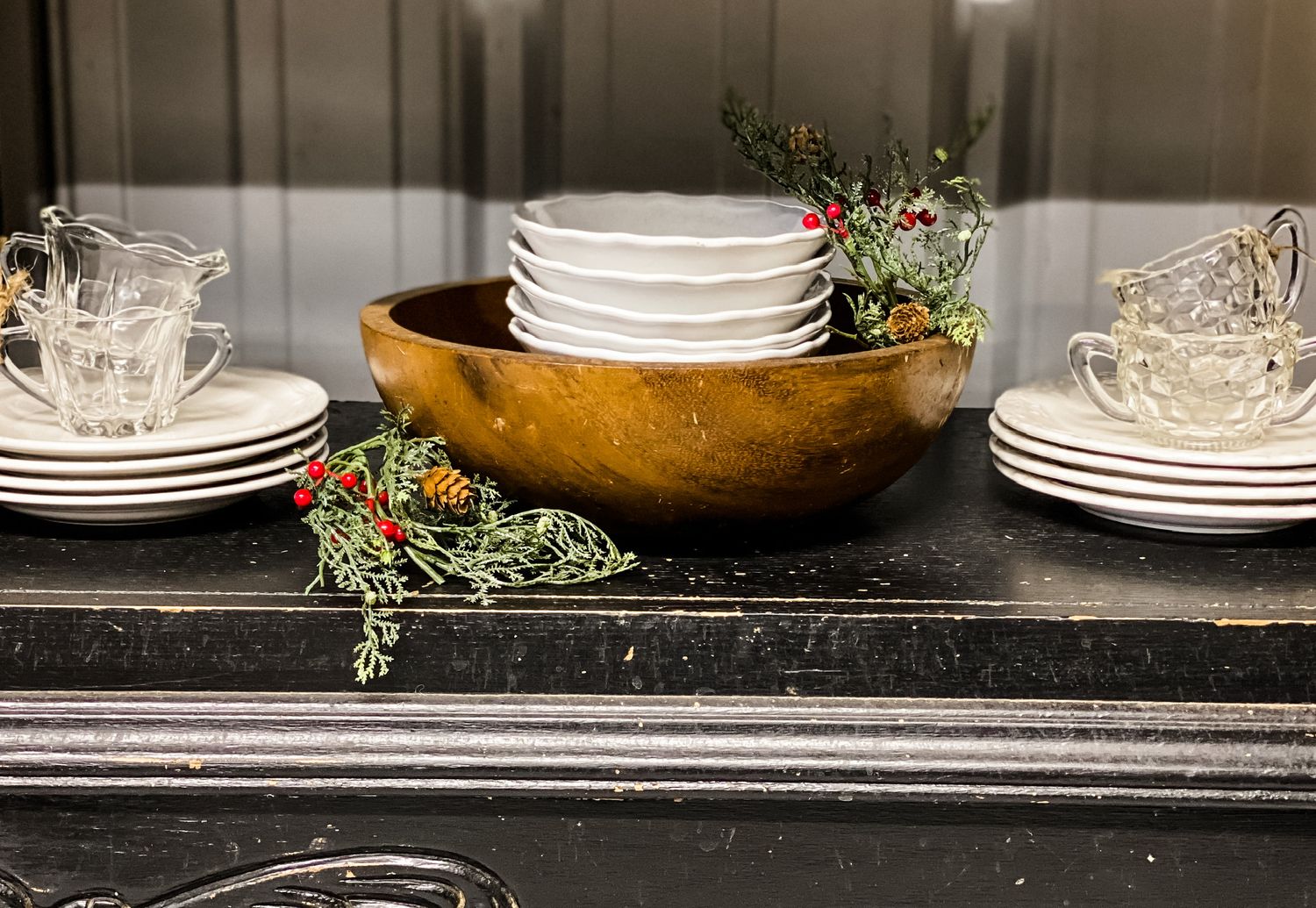 White plates and dishes arranged in wooden bowl with holiday greenery on dark marble counter.