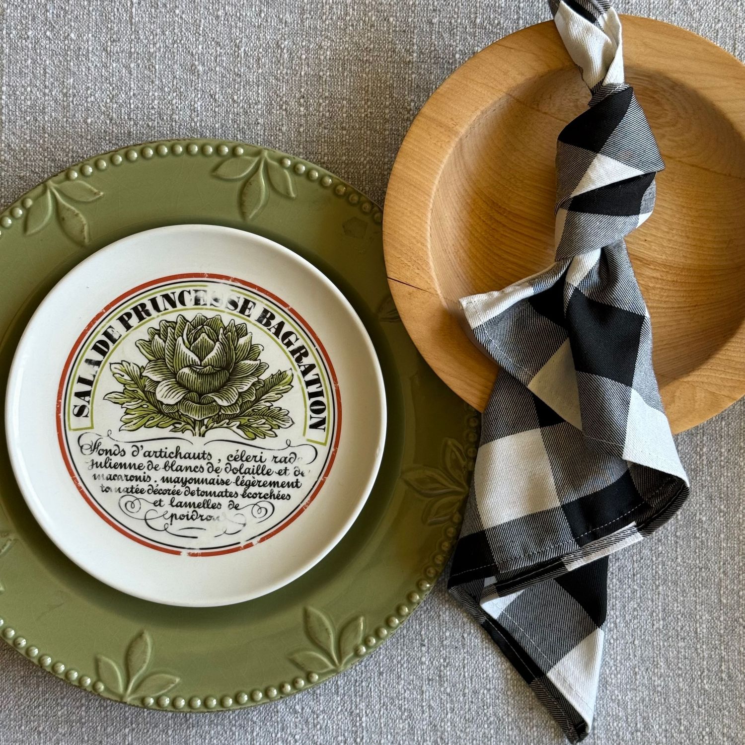 Green embossed plate with French salad recipe design, wooden charger, and black and white buffalo check napkin.