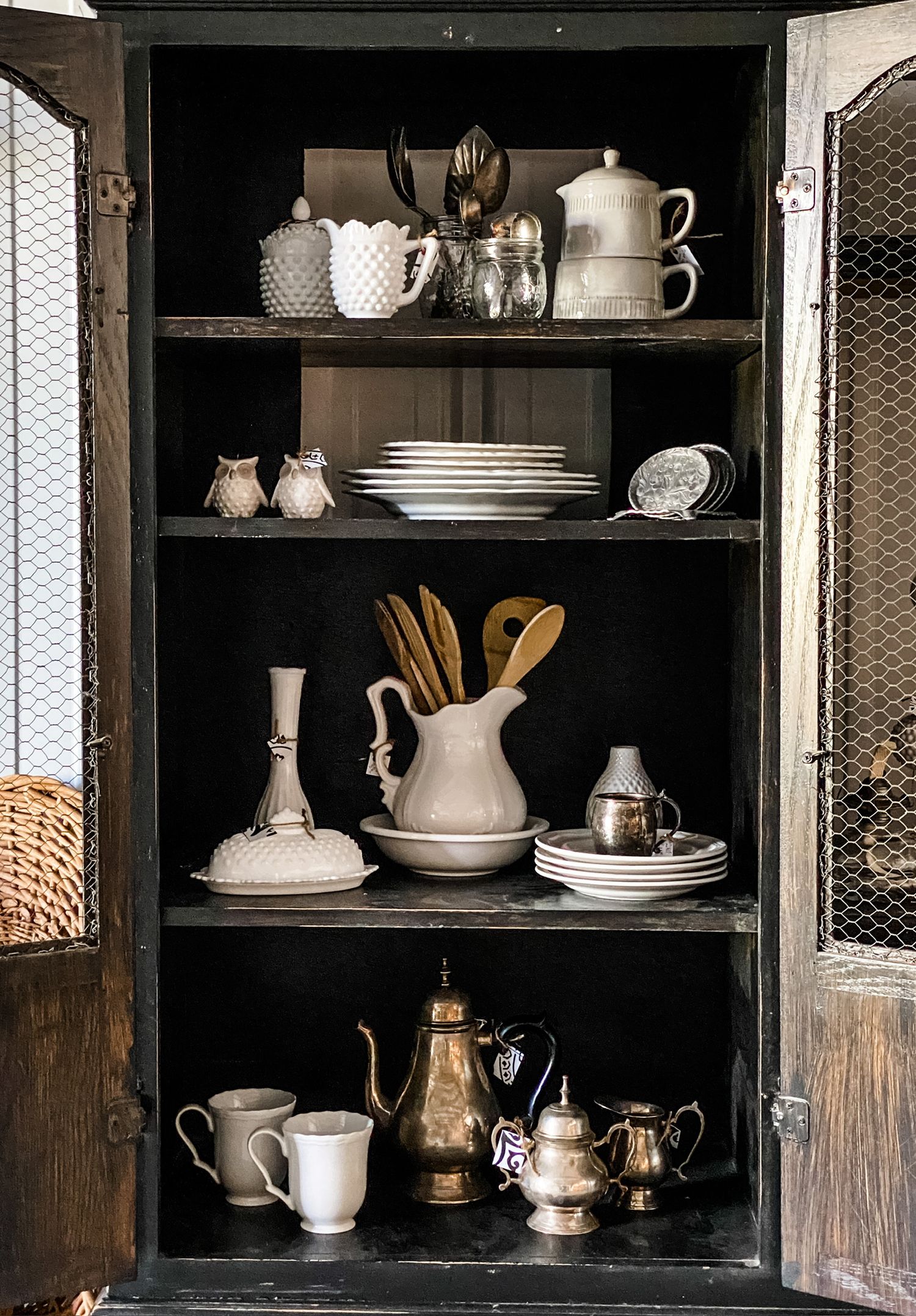 Vintage hutch displaying white ceramic dishes, teapots and wooden utensils on black shelves.