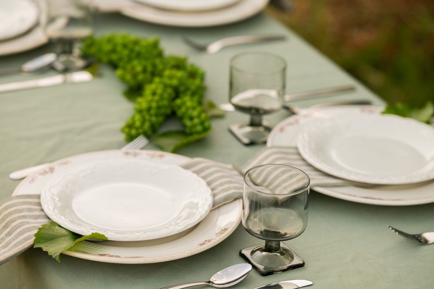 Elegant outdoor table setting with white plates, striped napkins, glass tumblers, and green botanical centerpiece on sage linen.