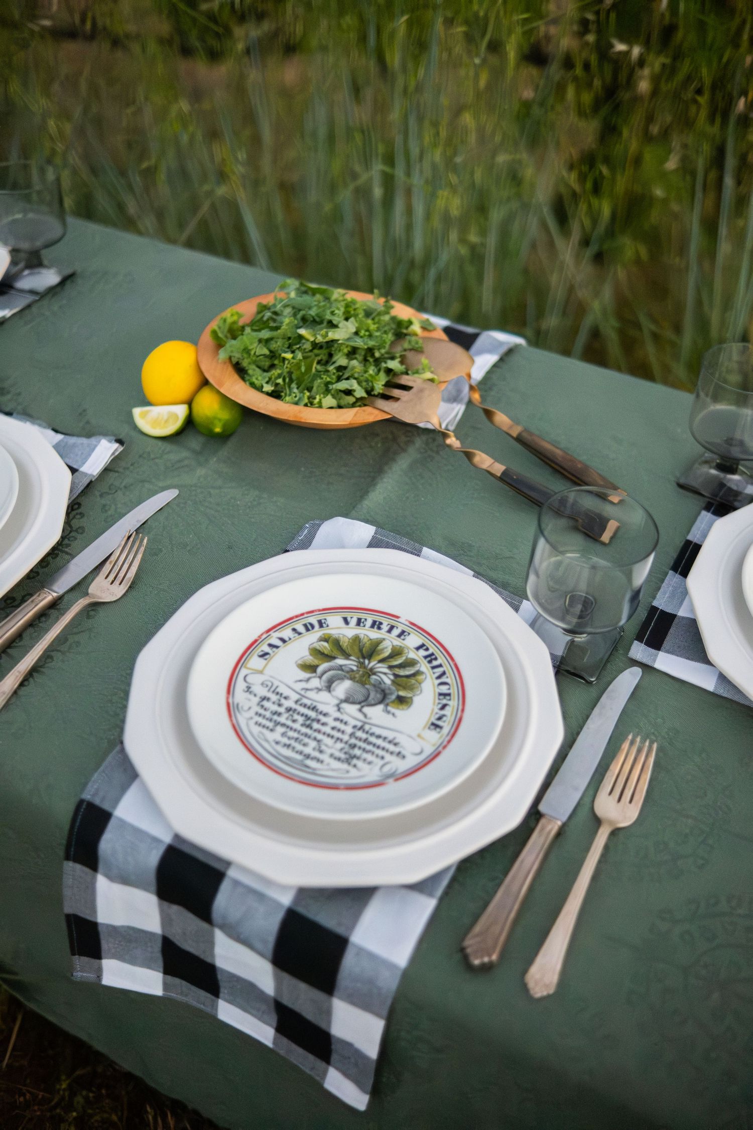 Outdoor table setting with green tablecloth, black gingham napkins, decorative plates, silverware, and fresh herbs in wooden bowl.