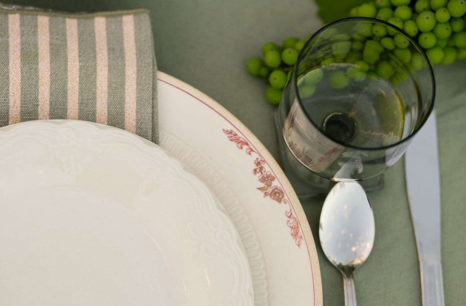 Elegant table setting with white floral plate, striped napkin, silver spoon, wine glass, and green grapes on green cloth.