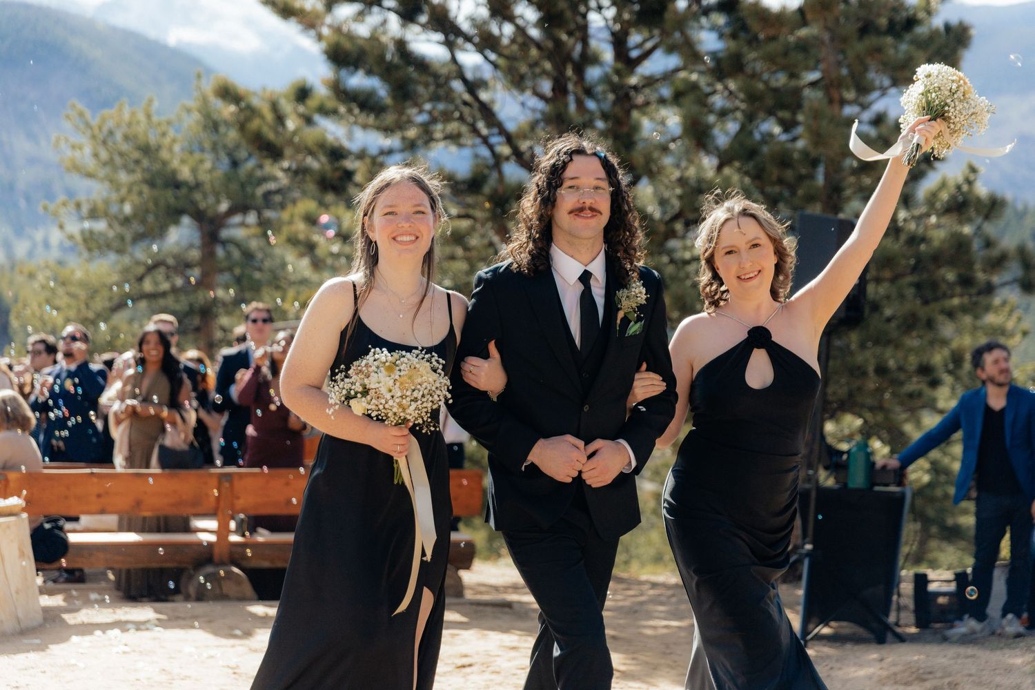 Two bridesmaids in black dresses walk with groomsman at outdoor mountain wedding ceremony.