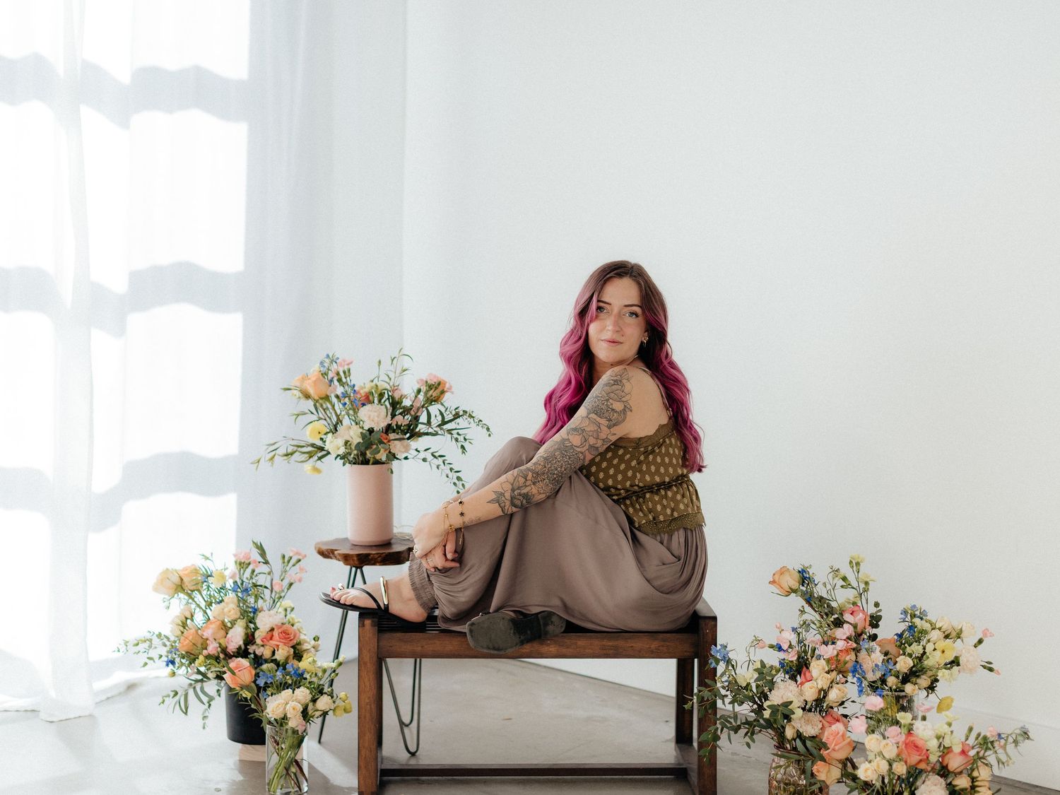 Woman with pink hair sits on a wooden bench surrounded by colorful floral arrangements in a bright white studio.