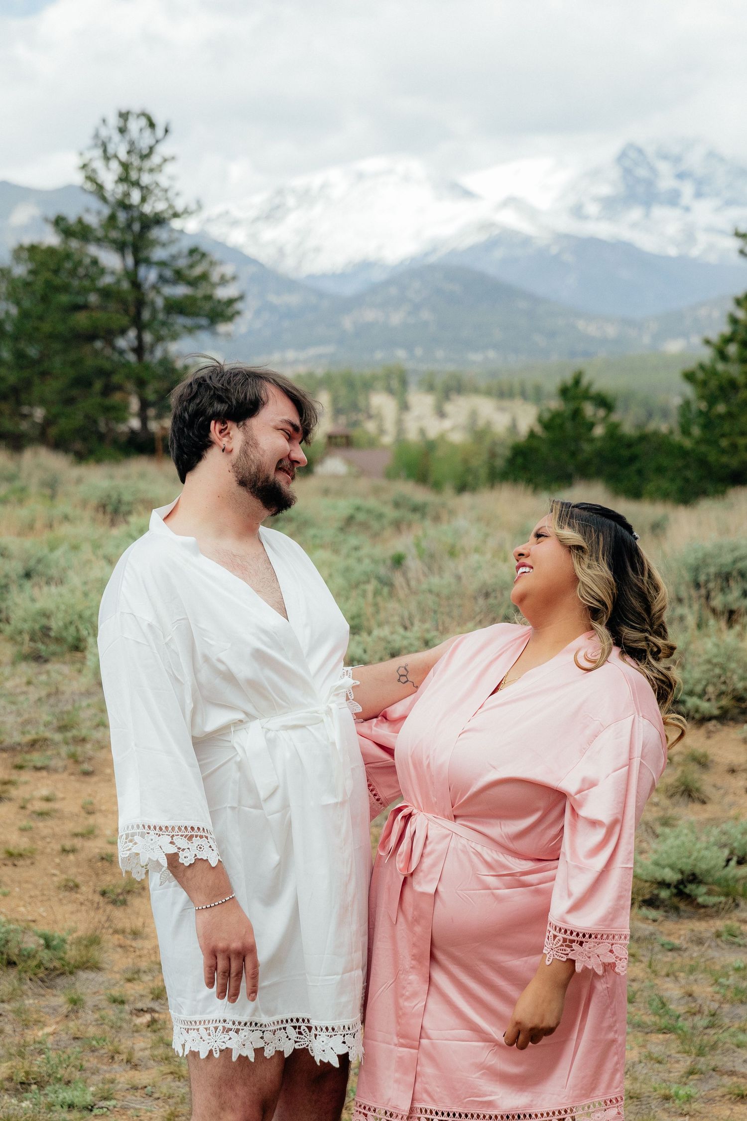 Couple in matching robes smiling at each other with snow-capped Rocky Mountains in the background.
