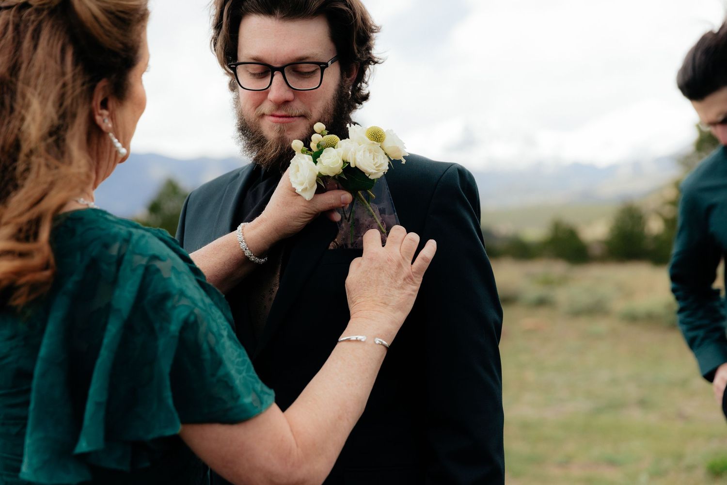 Woman pins white boutonniere on groom's black suit lapel at outdoor mountain wedding.