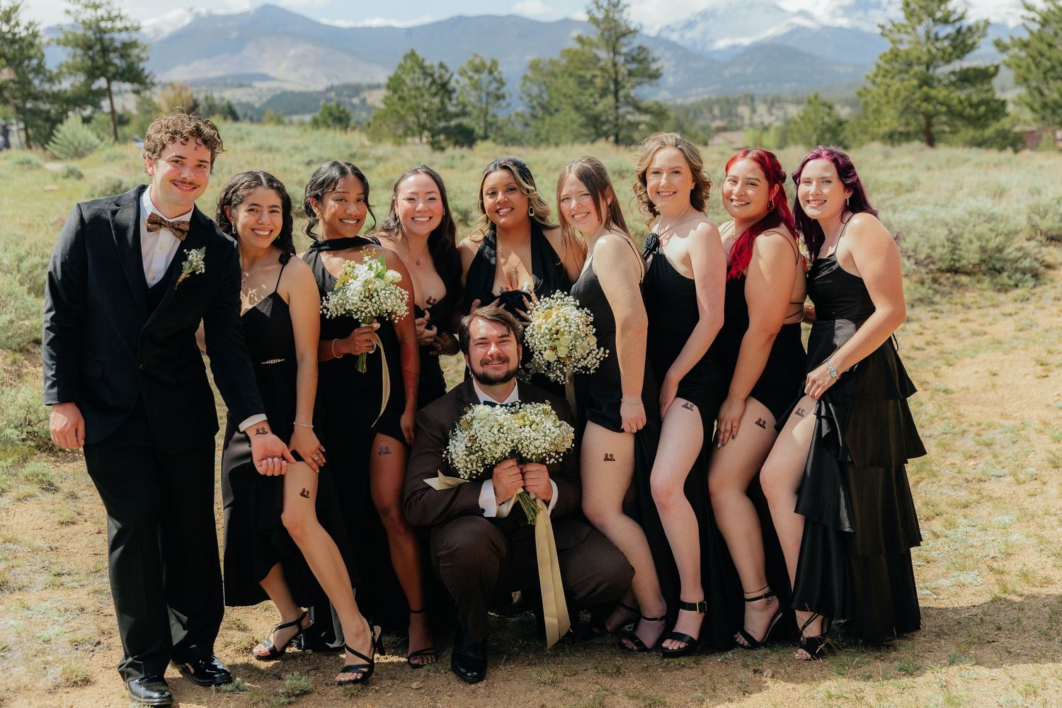 Groom poses with wedding party of bridesmaids in black dresses with mountain backdrop.