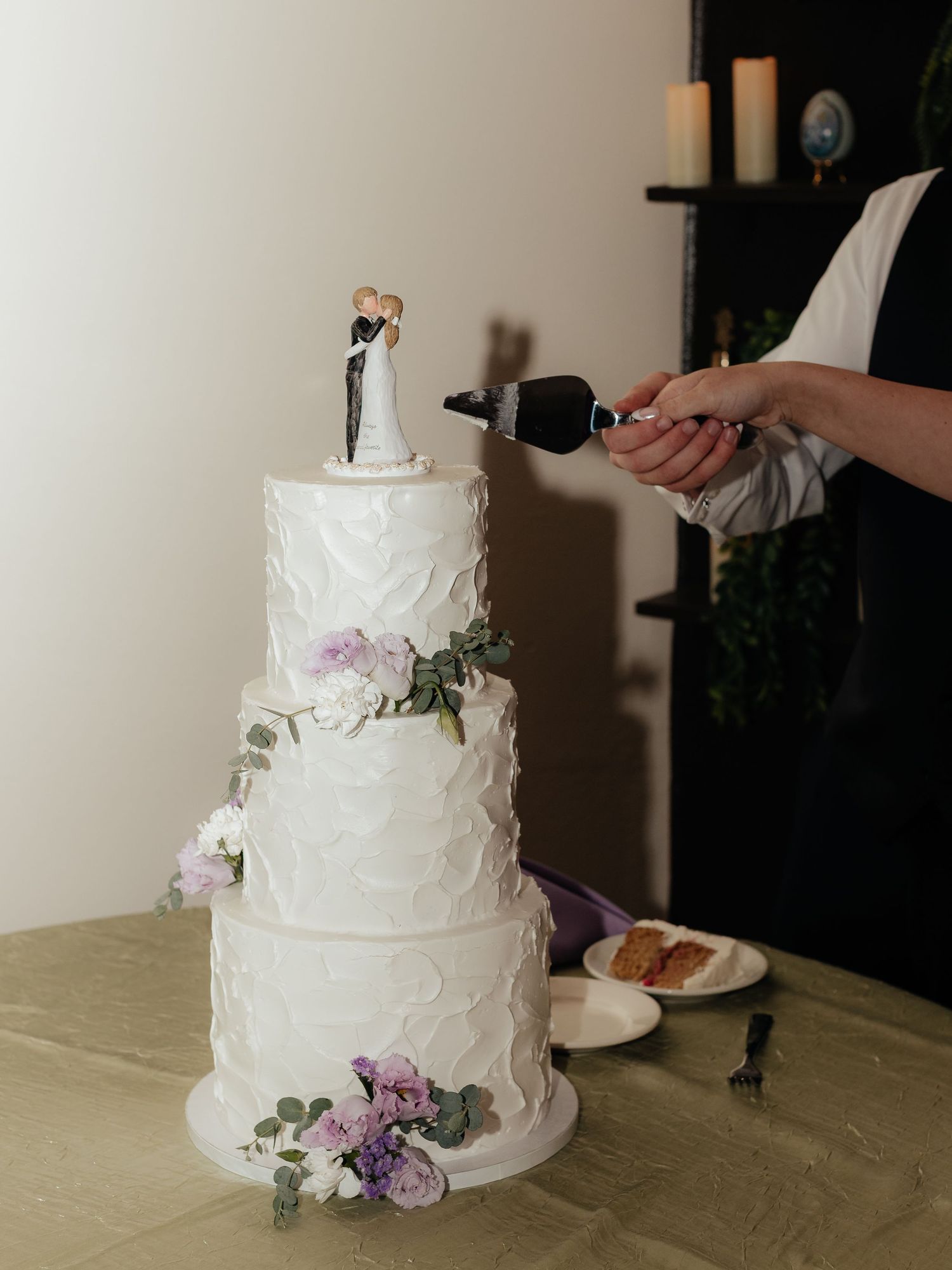 Four-tier white wedding cake with floral decorations and bride and groom topper being cut at reception.