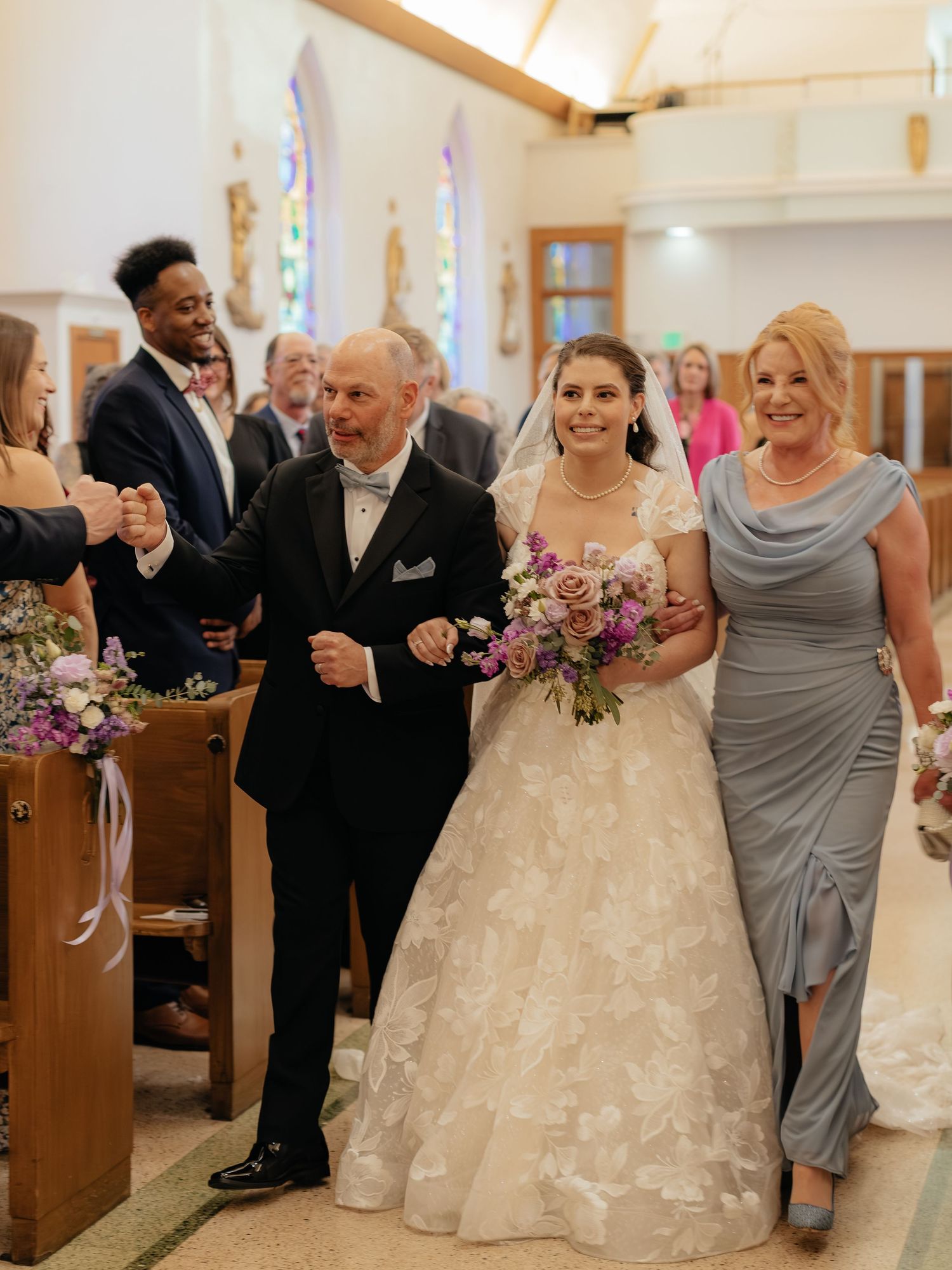 Bride walks down church aisle with parents, holding purple bouquet in elegant lace ball gown wedding dress.