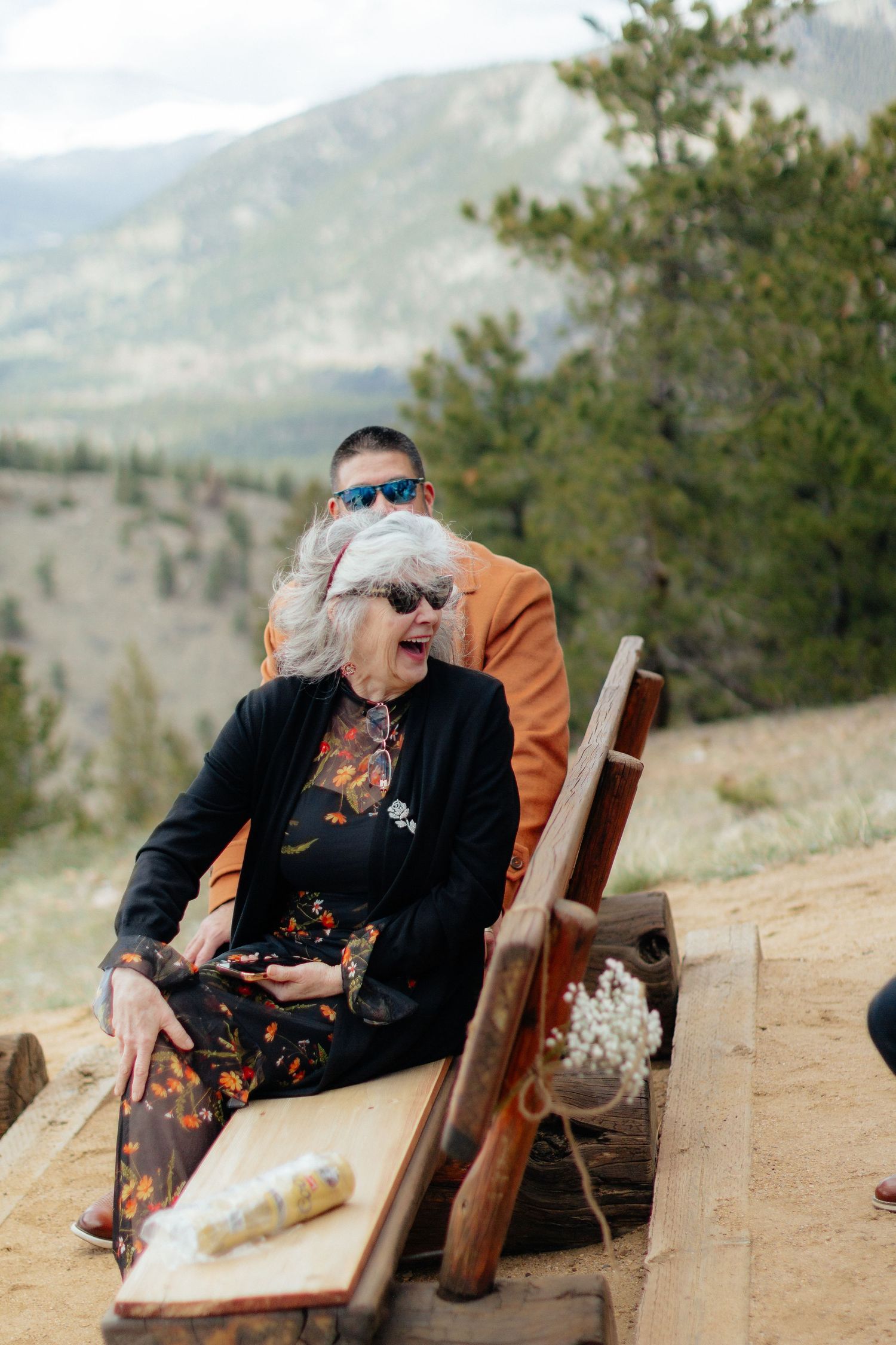 Elderly woman in black jacket and sunglasses seated on rustic wooden bench at mountain wedding ceremony.