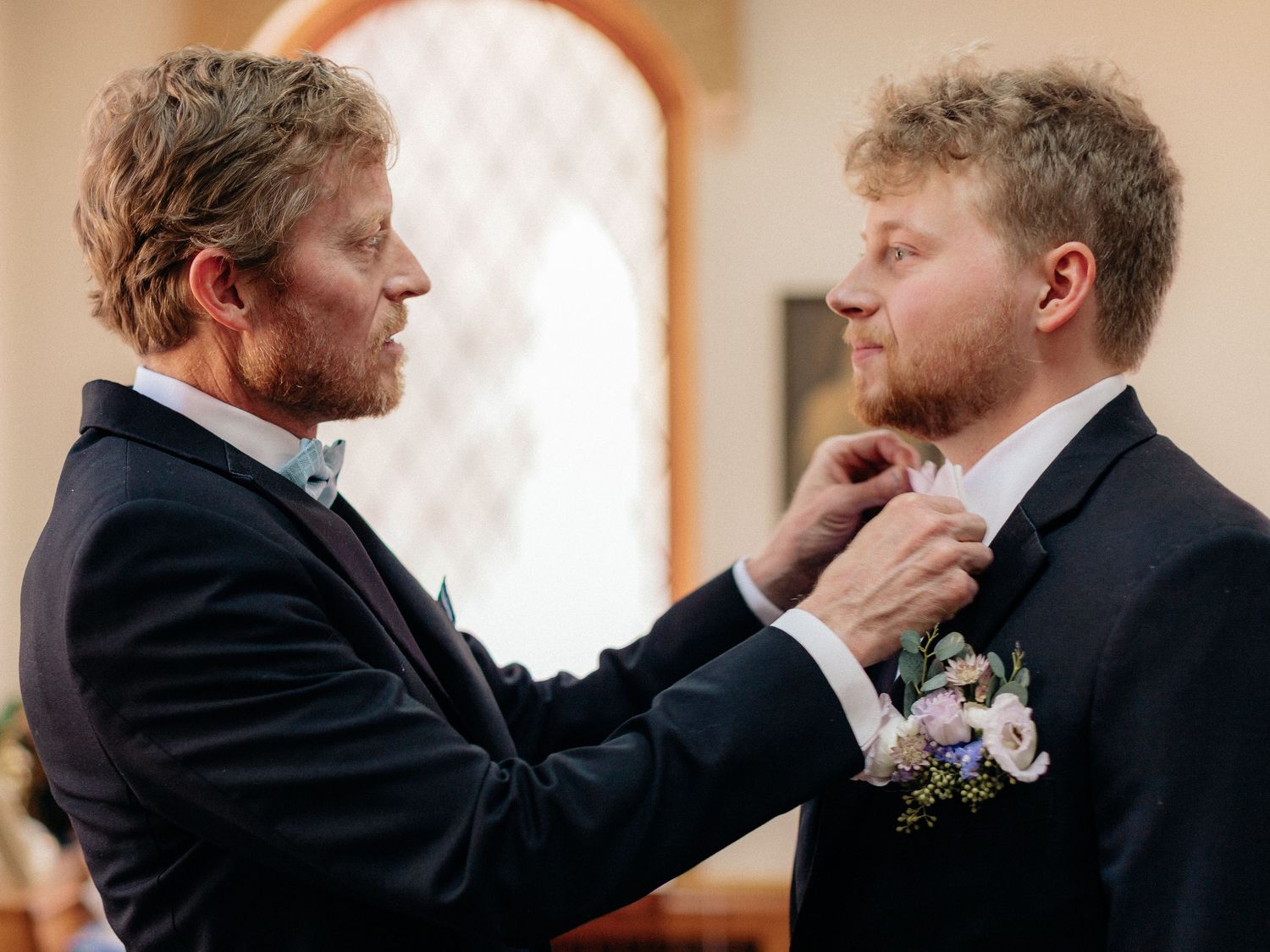 Two grooms in navy suits adjusting bow tie at wedding ceremony with floral boutonniere.