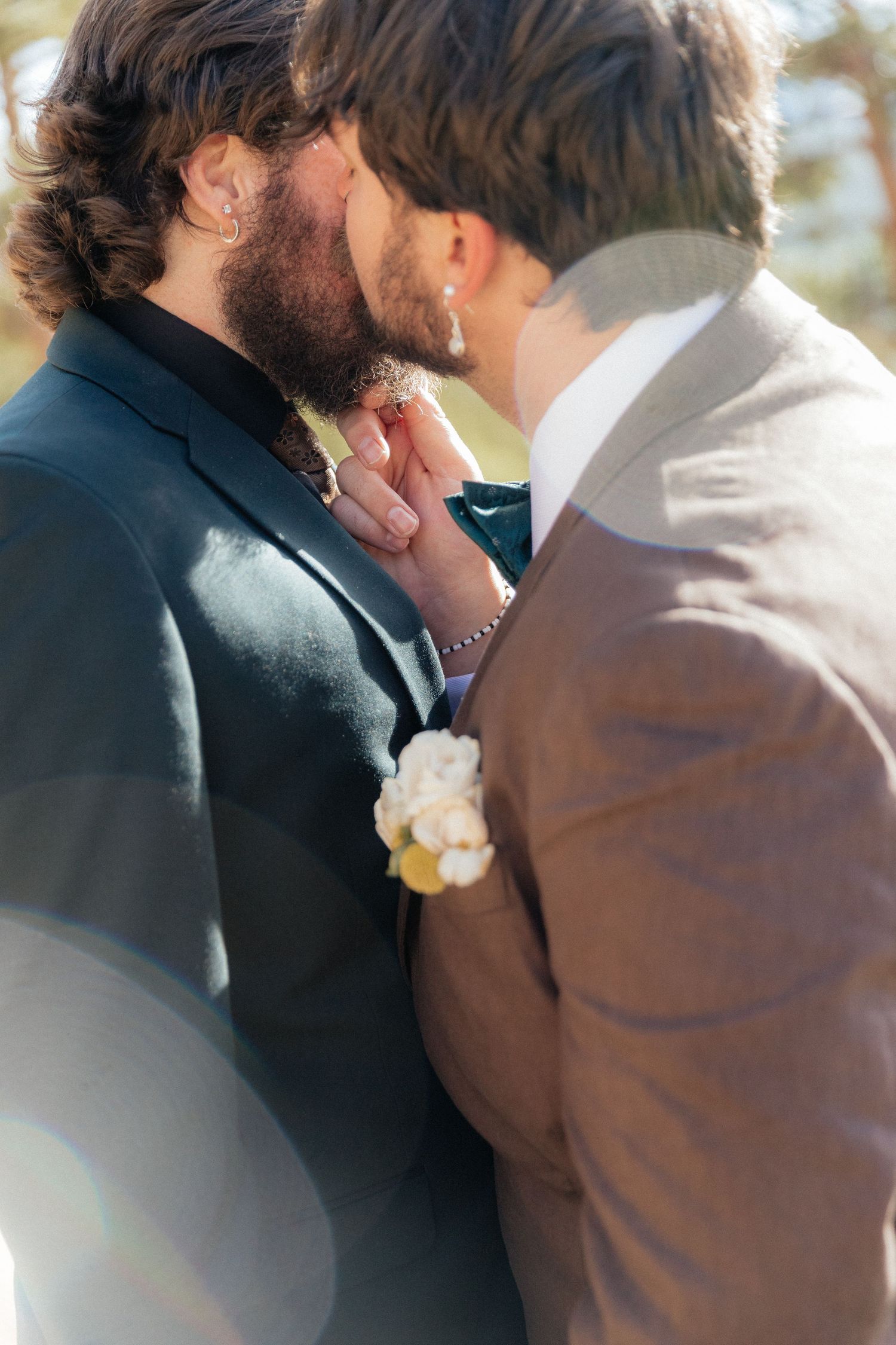 Two grooms in suits share an intimate moment, one wearing a white floral boutonniere, in a sunlit outdoor wedding setting.