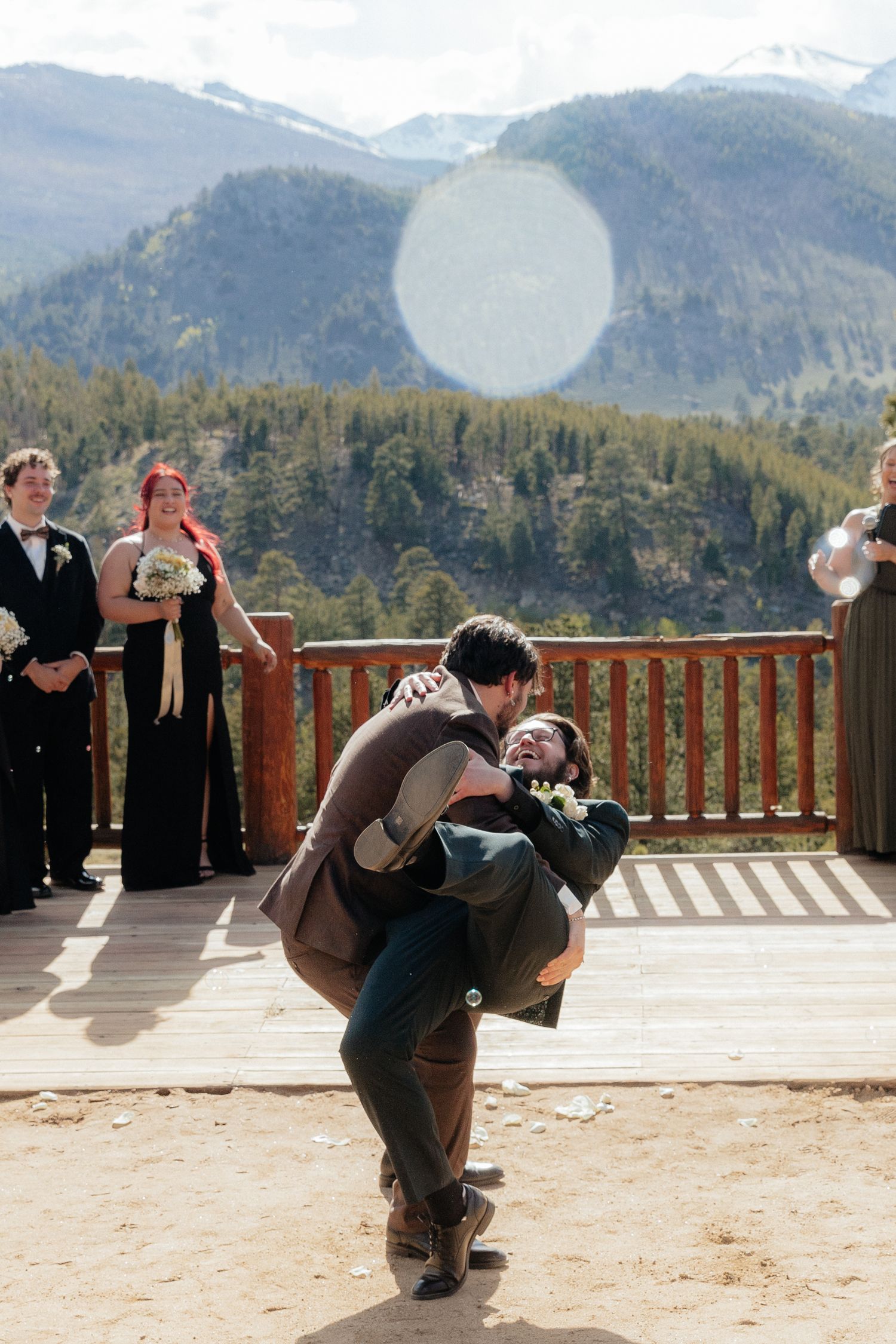 Groom lifting bride during outdoor mountain wedding ceremony on wooden deck with scenic Rocky Mountain backdrop.